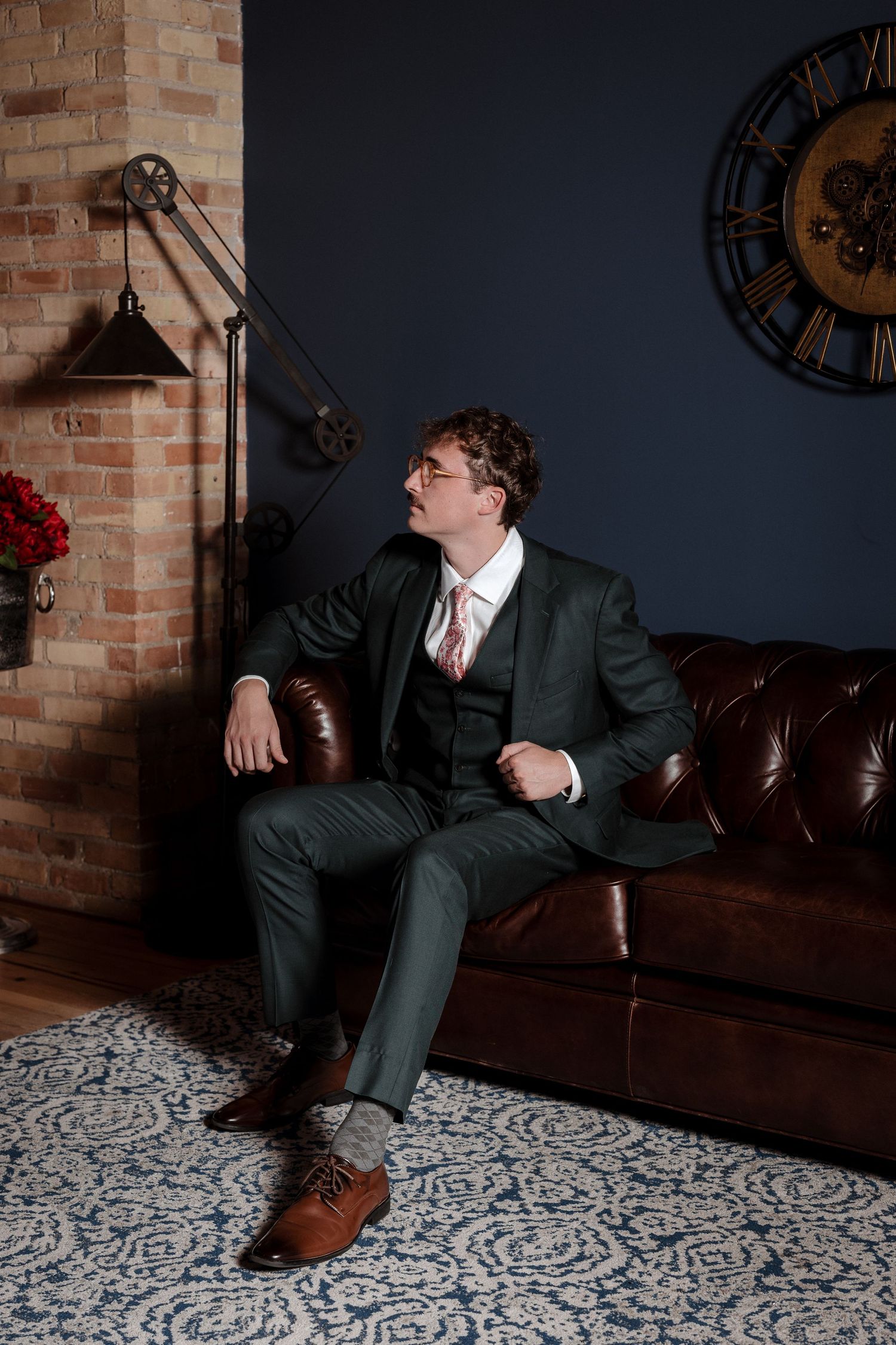 Dapper groom in dark three-piece suit sits on leather sofa in industrial-chic wedding venue interior.