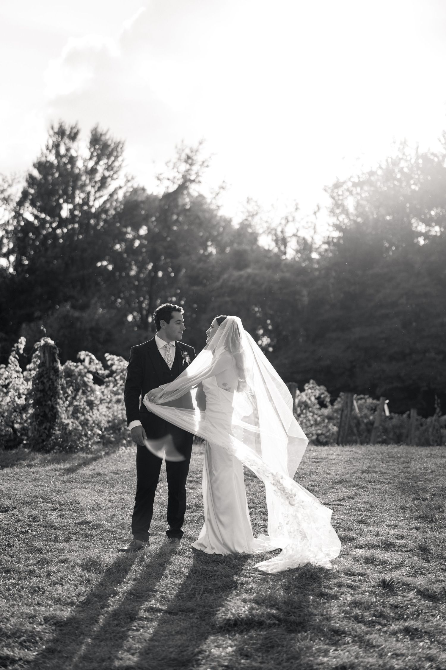 Black and white romantic photo of bride and groom standing together outdoors with veil flowing in wind.