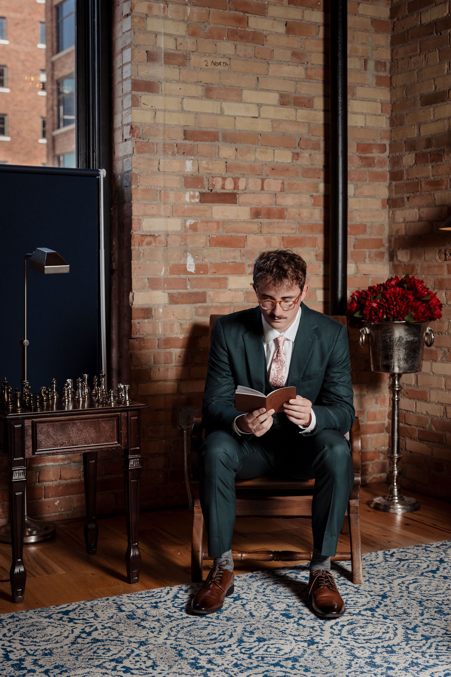 Groom in a dark suit reading vows from a small book while seated in a brick-walled room.