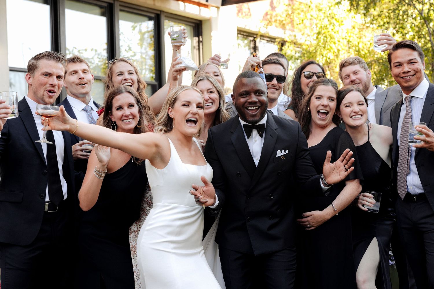 Joyful wedding party group photo with bride in white dress and groom in tuxedo surrounded by smiling guests outdoors.