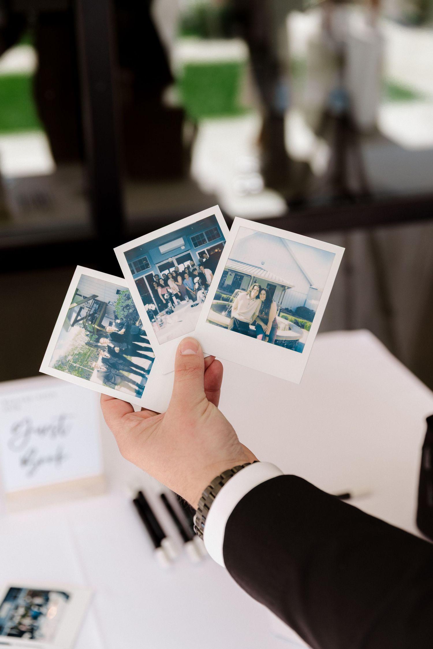 A hand holding three Polaroid photos near a guest book table at a wedding reception outdoor event.