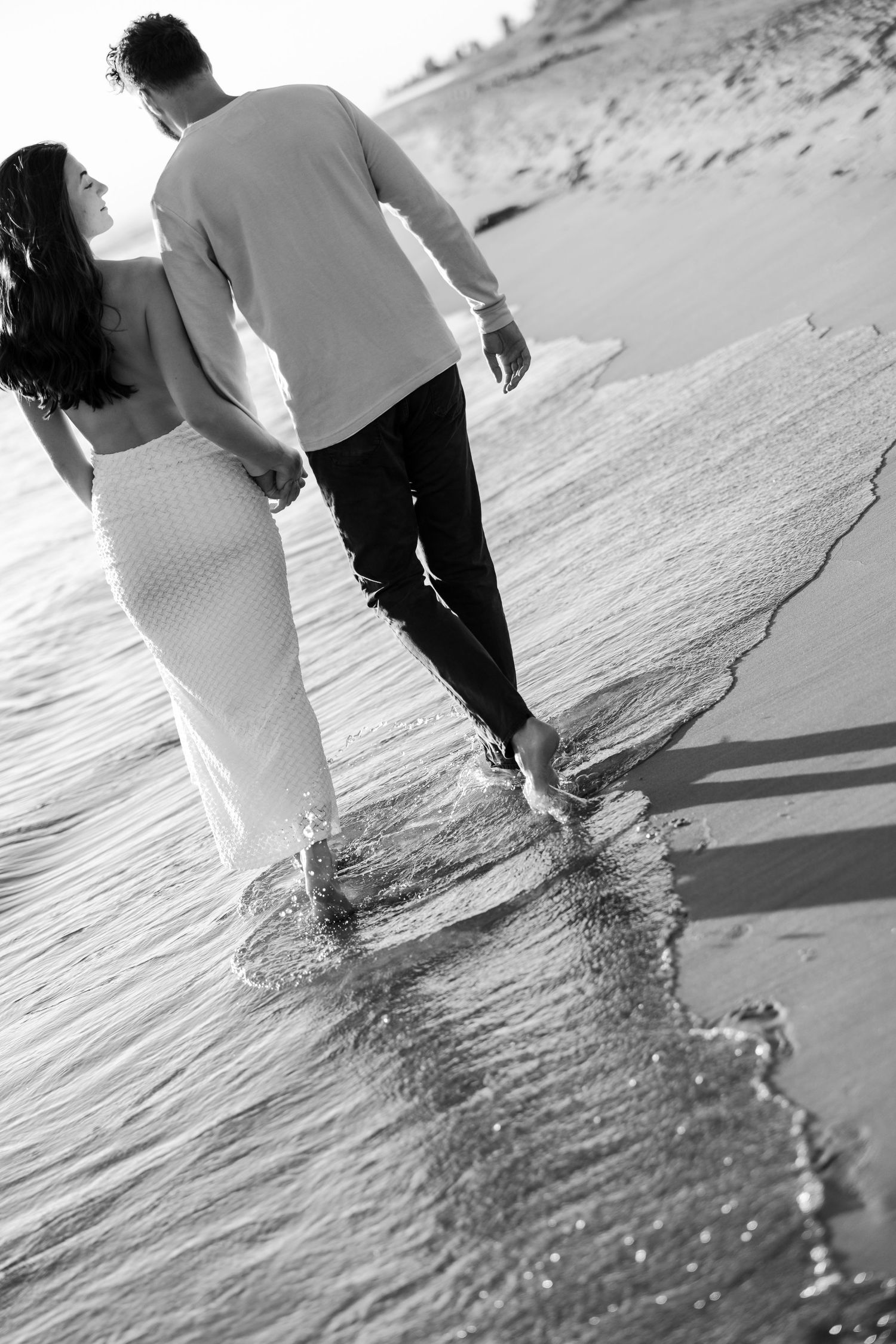 Black and white photo of a couple holding hands walking barefoot along a sandy beach shoreline.