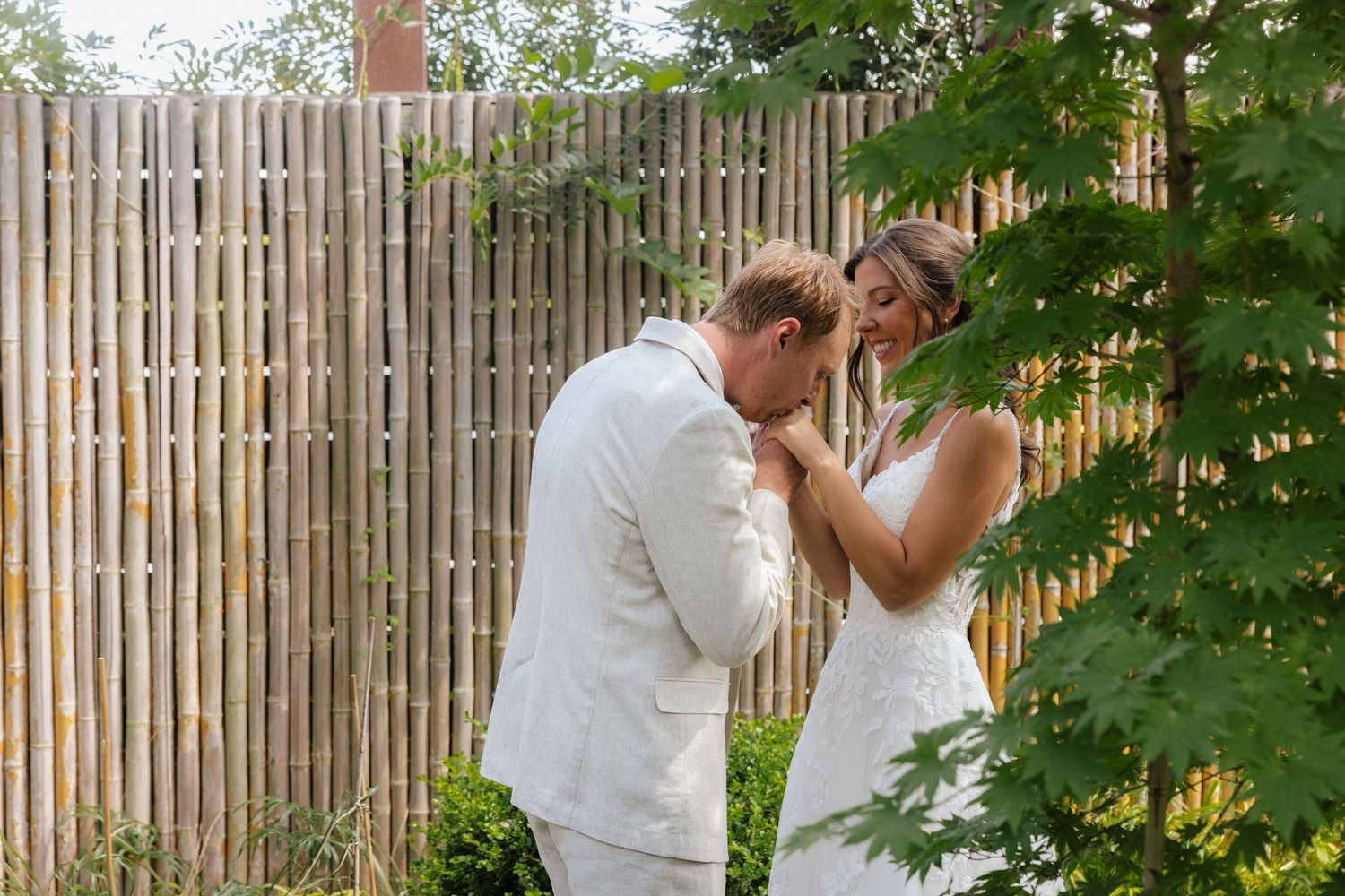 Groom in white suit kissing bride's hand near bamboo fence in romantic outdoor wedding ceremony.