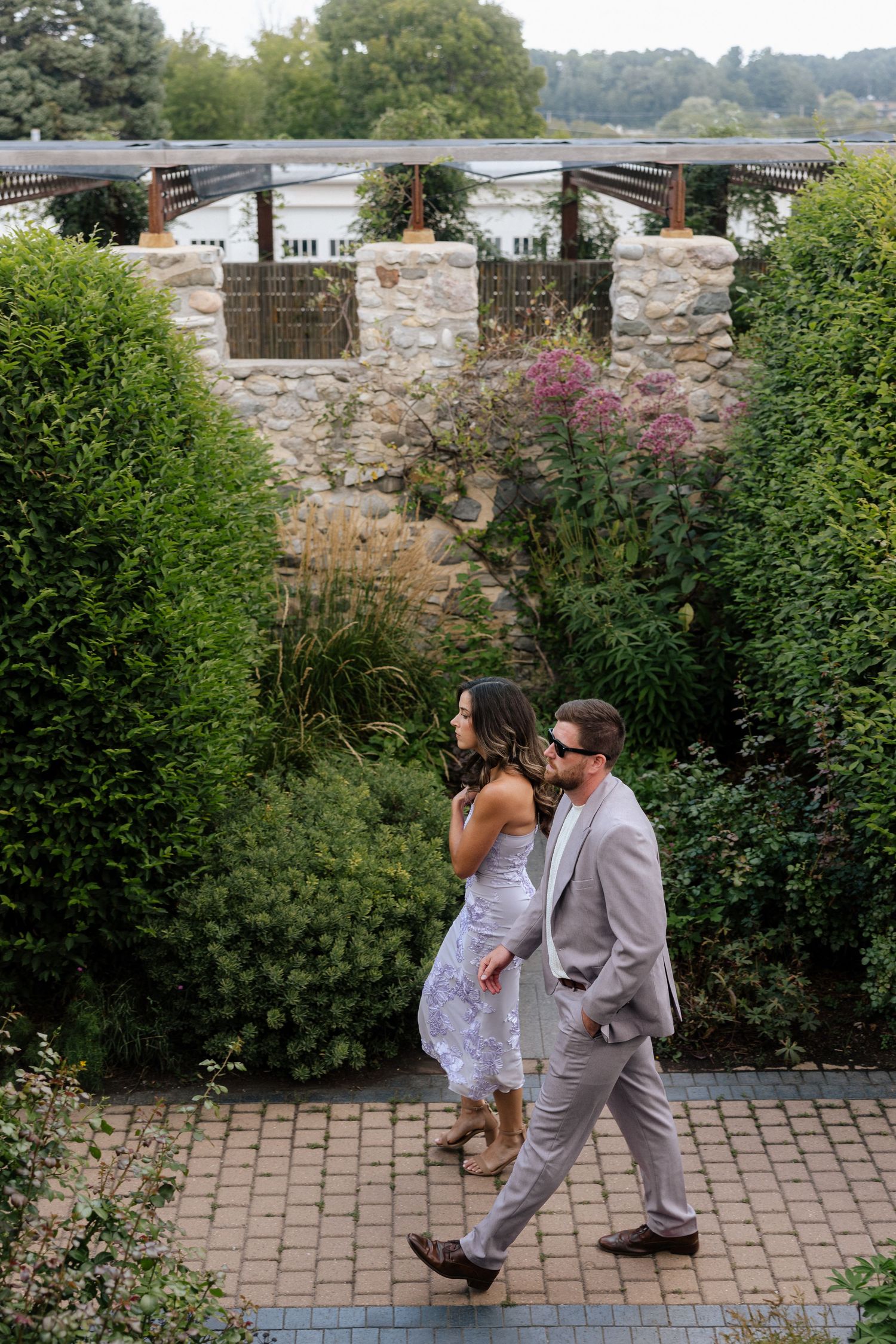 Couple in formal attire walking through a lush garden with stone wall and pergola backdrop.