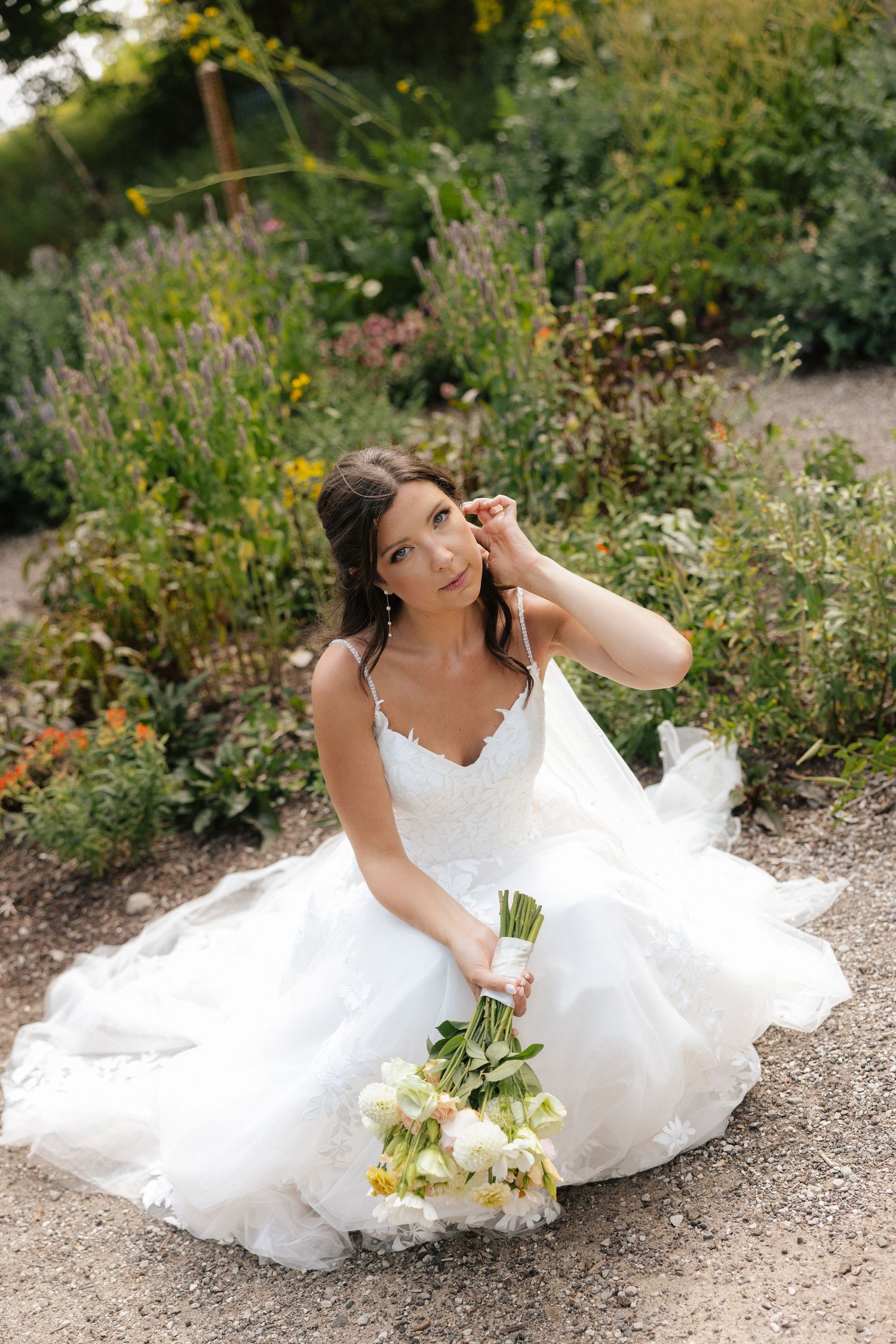 Bride in a flowing white lace wedding dress sits on a garden path holding a bouquet of white and yellow flowers.