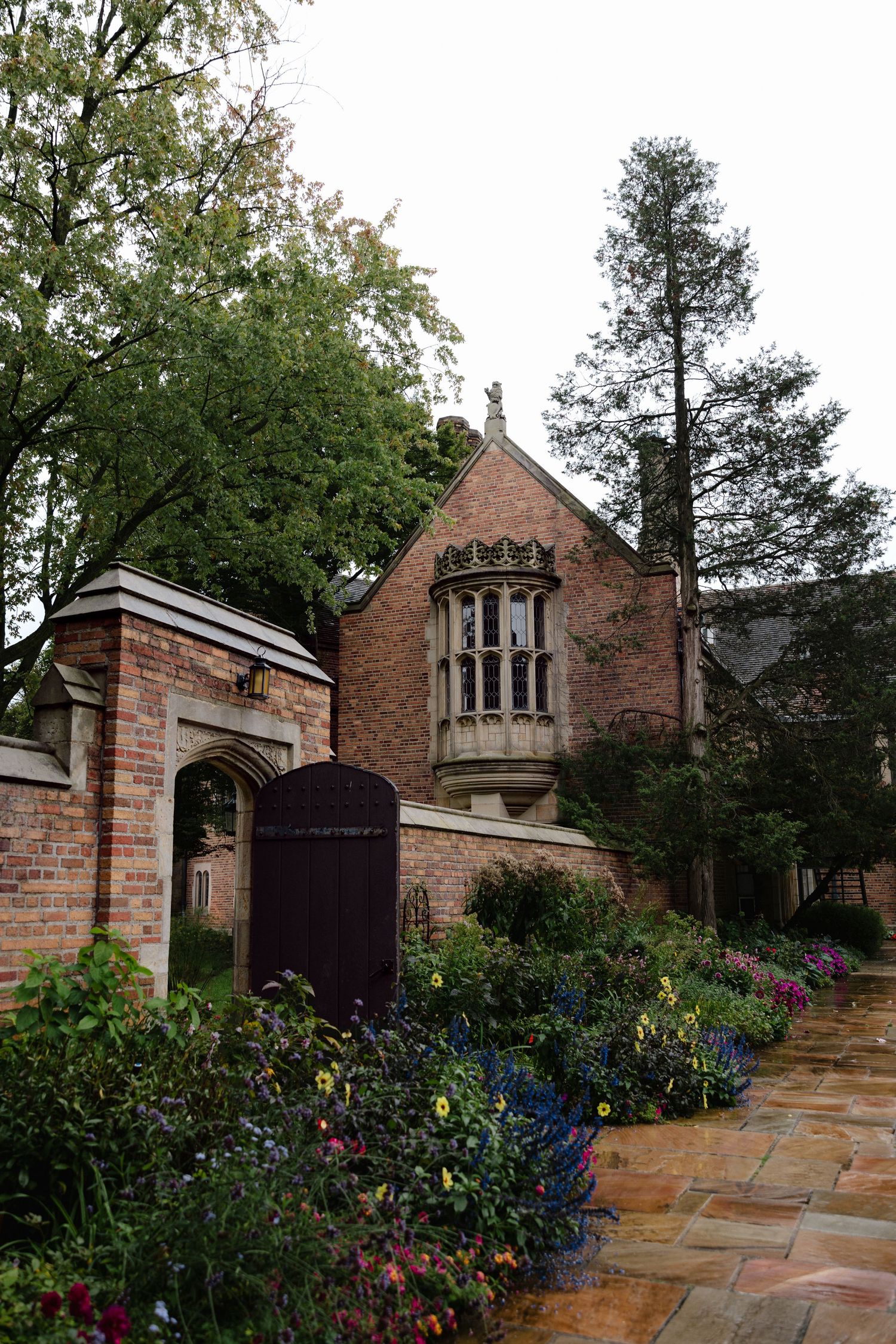 Gothic brick mansion with ornate bay window, colorful flower garden, and arched gate along a stone pathway.