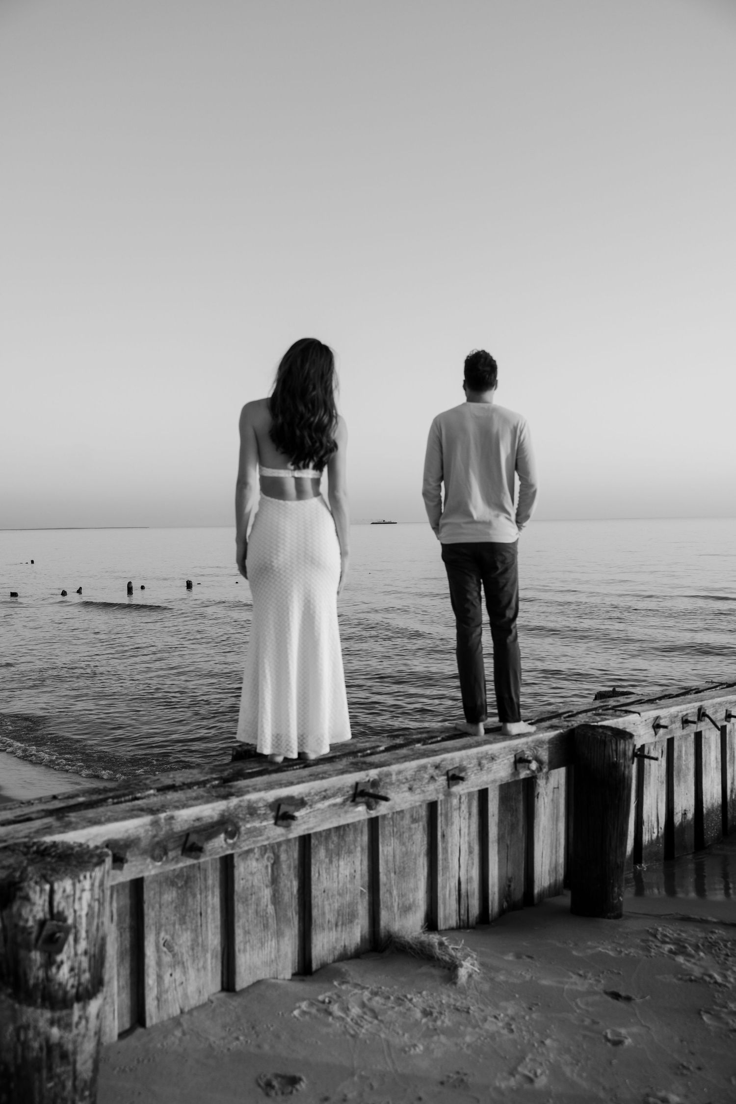 Black and white photo of a couple standing apart on a seaside pier, gazing at the ocean horizon.