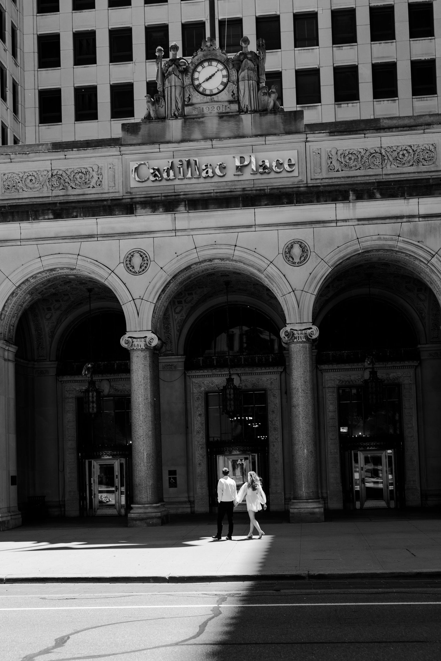 Black and white photo of a couple walking in front of Cadillac Place historic building with arched entrance in Detroit.