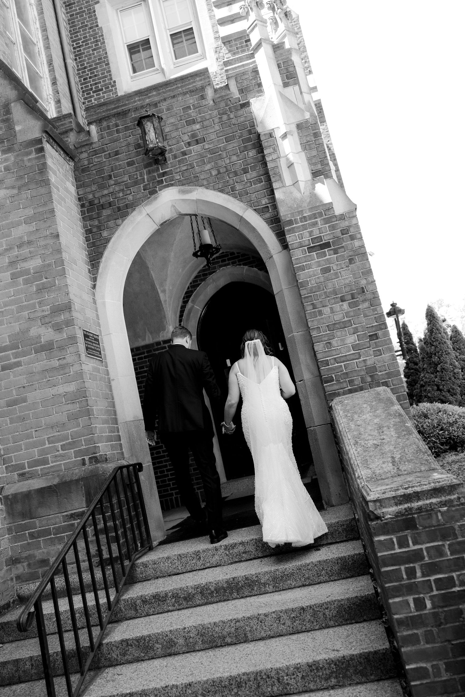 Black and white photo of bride and groom holding hands walking up stone steps into arched church doorway.