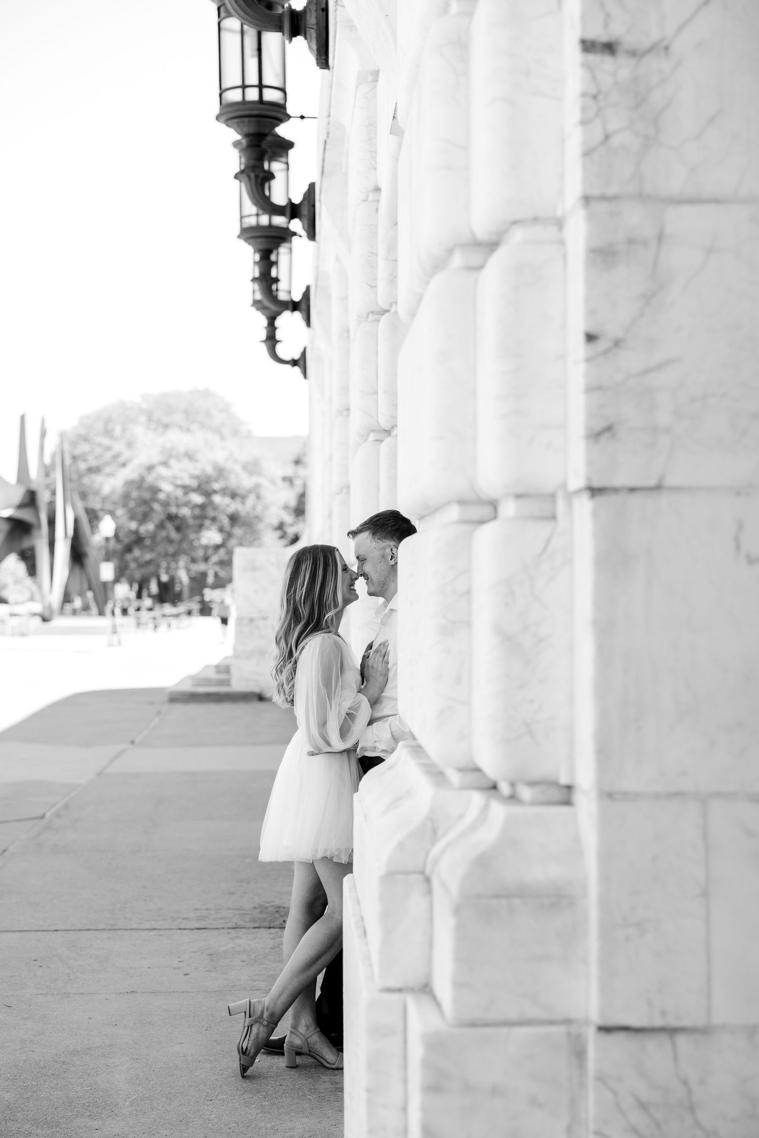 Black and white photo of a couple sharing an intimate moment leaning against ornate stone columns on a city sidewalk.
