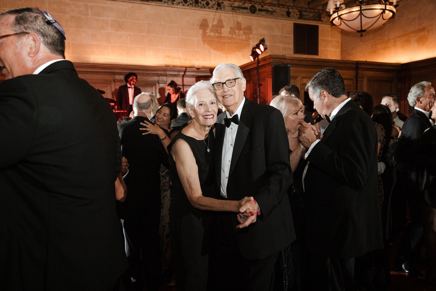 Elegant couple dancing at a formal black-tie gala event in a grand ballroom with other guests.