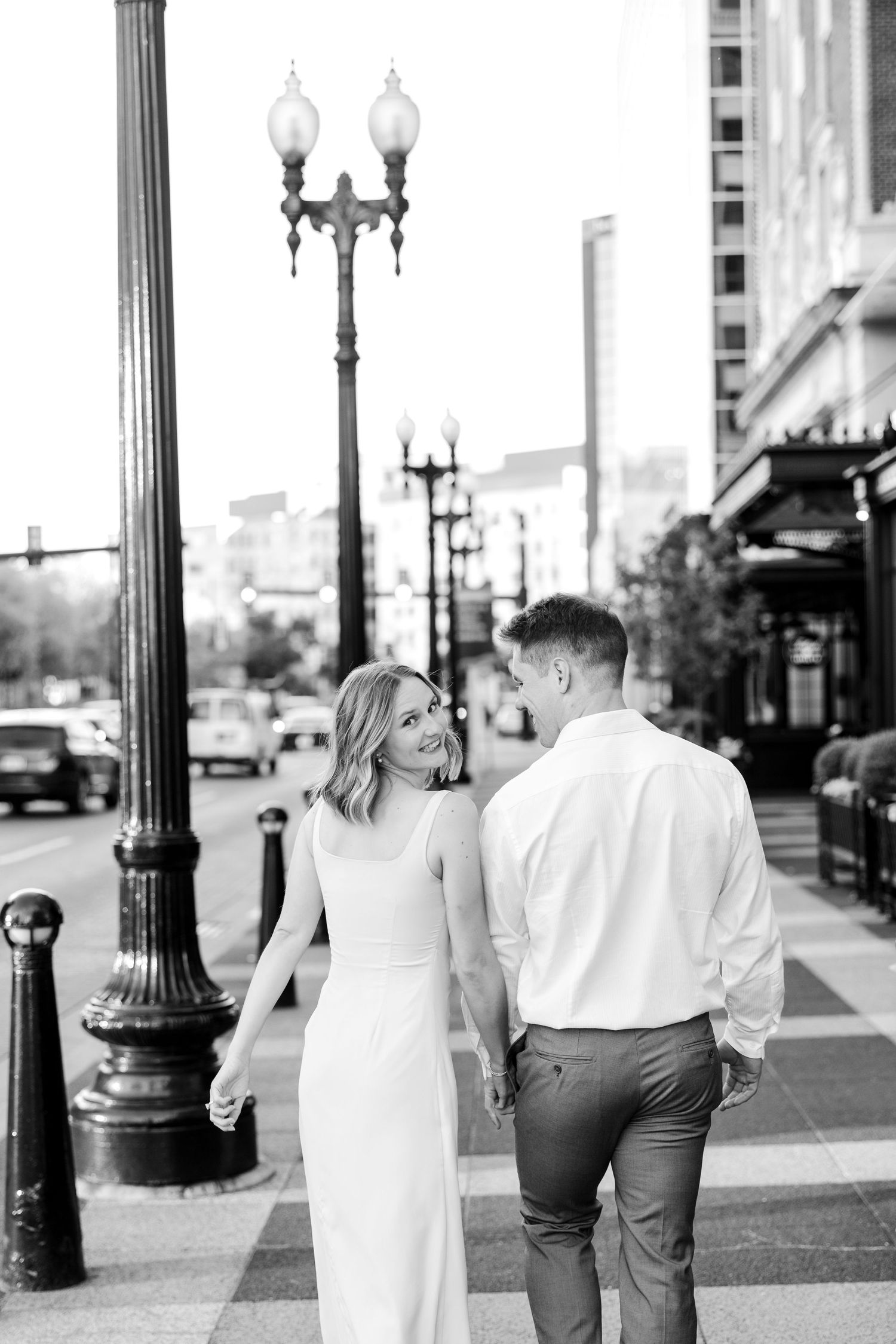 Black and white photo of a couple holding hands walking on a city sidewalk, woman in white dress smiling at man.