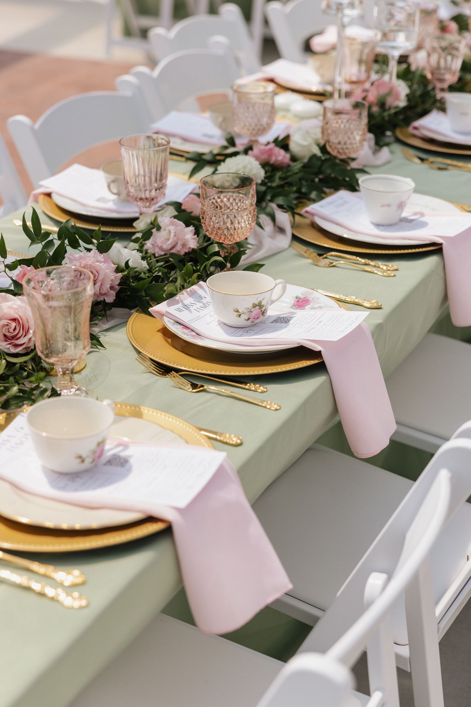 Elegant wedding table setting with sage green runner, pink roses, gold chargers, floral teacups, and pink napkins.