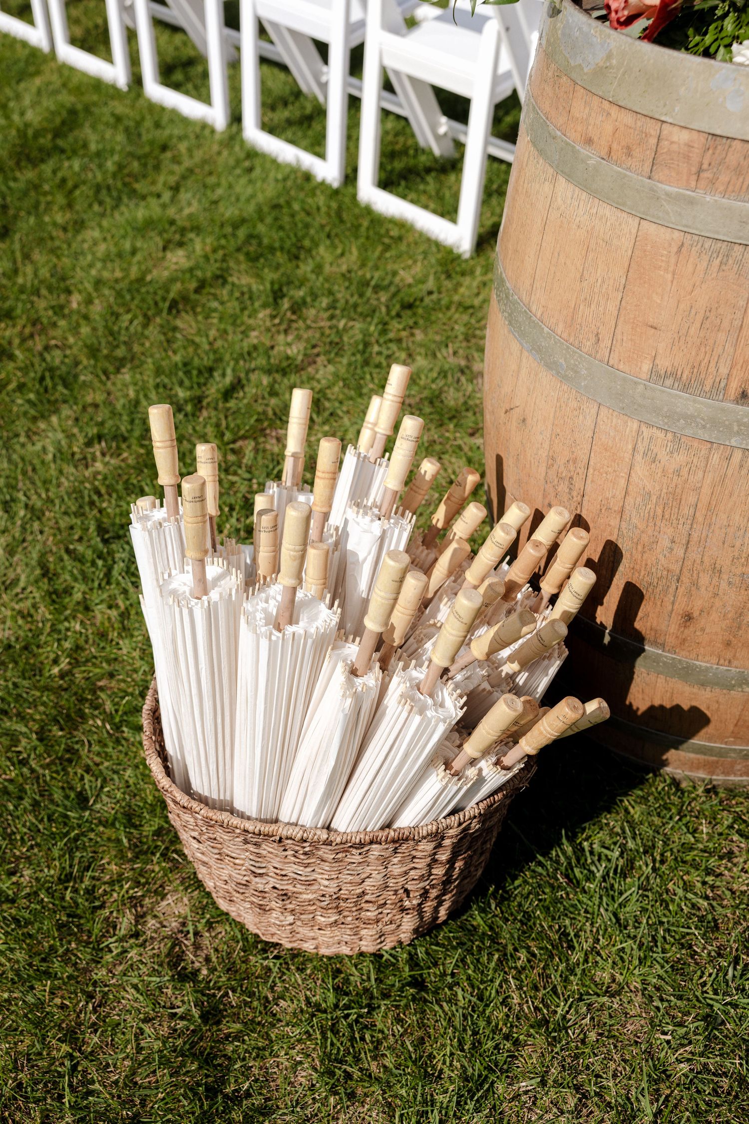 Basket of white parasol umbrellas beside wooden wine barrel at outdoor vineyard wedding ceremony setup.