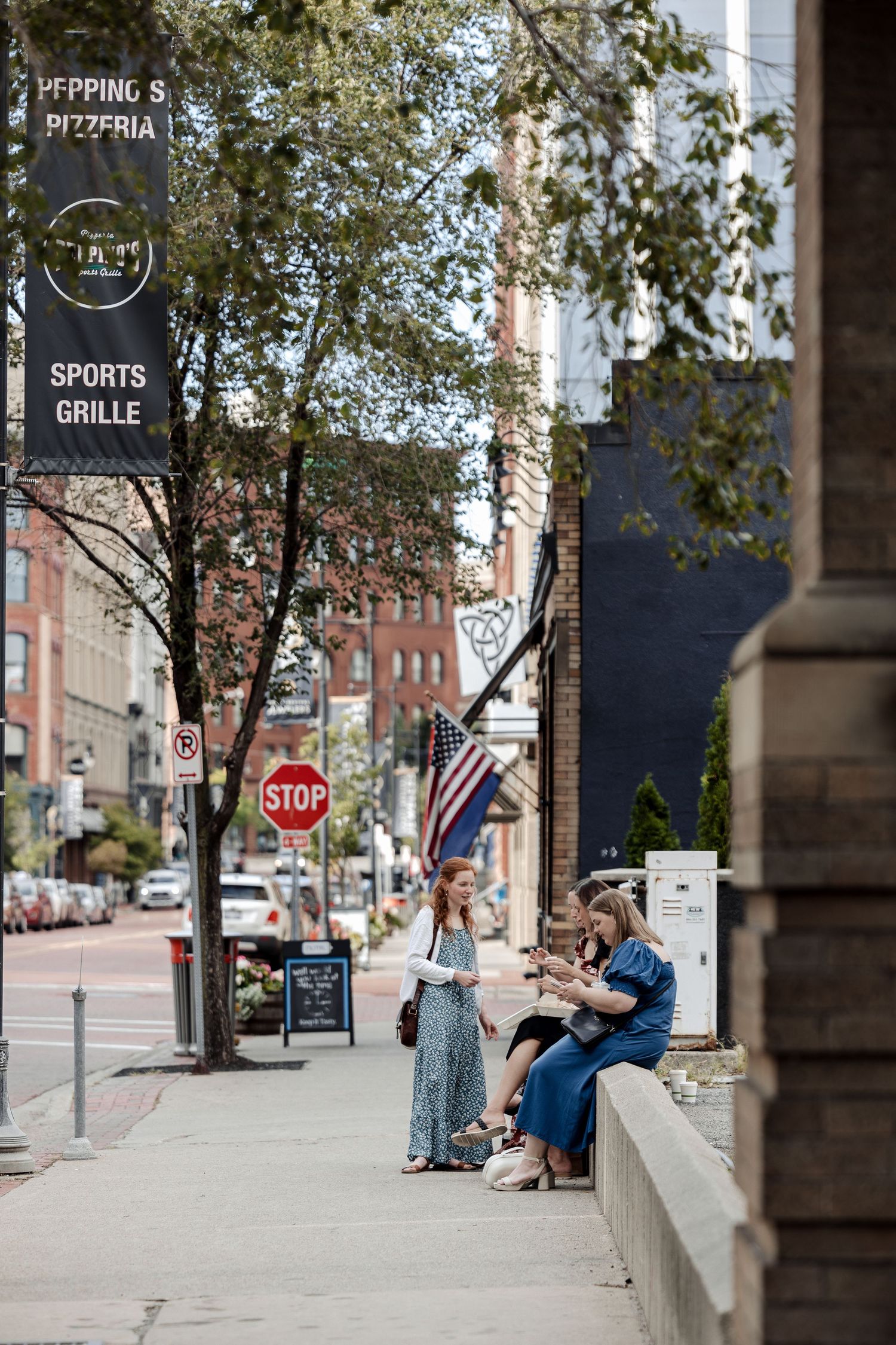 Three women wait on a city sidewalk near a pizzeria and sports grille with an American flag visible.
