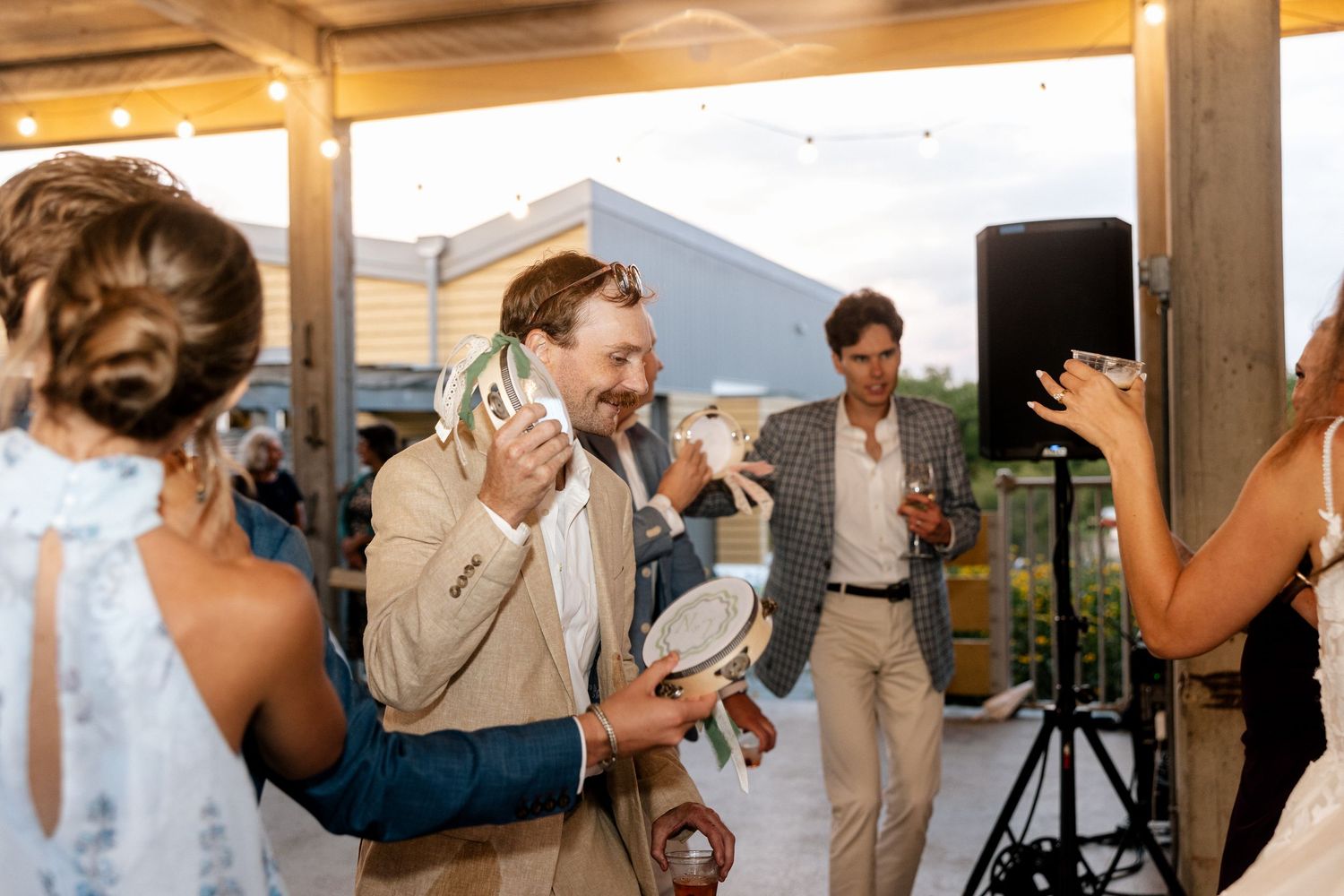 A man in a tan suit laughs and dances at an outdoor wedding reception while guests celebrate around him.