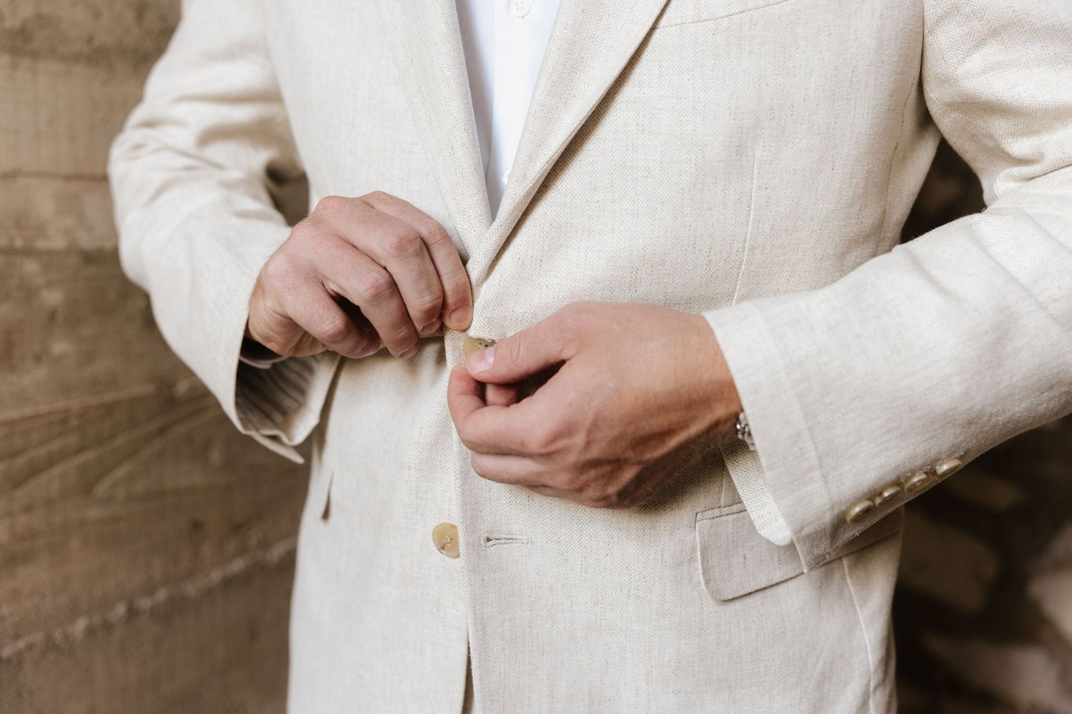 Man in beige linen suit buttoning jacket, close-up of hands fastening button on elegant cream blazer.