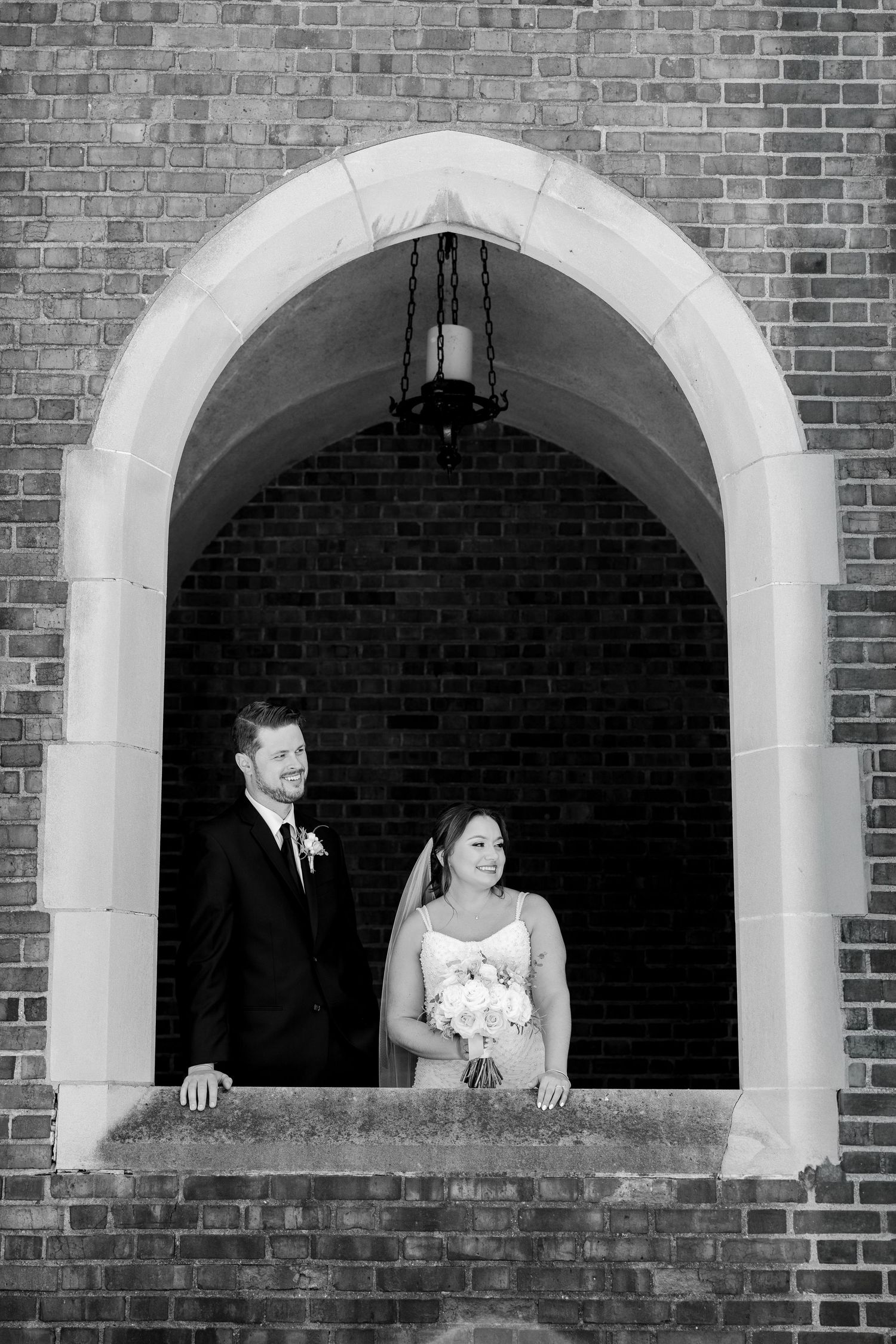 Black and white wedding portrait of bride and groom standing in a stone arch doorway of a brick church.