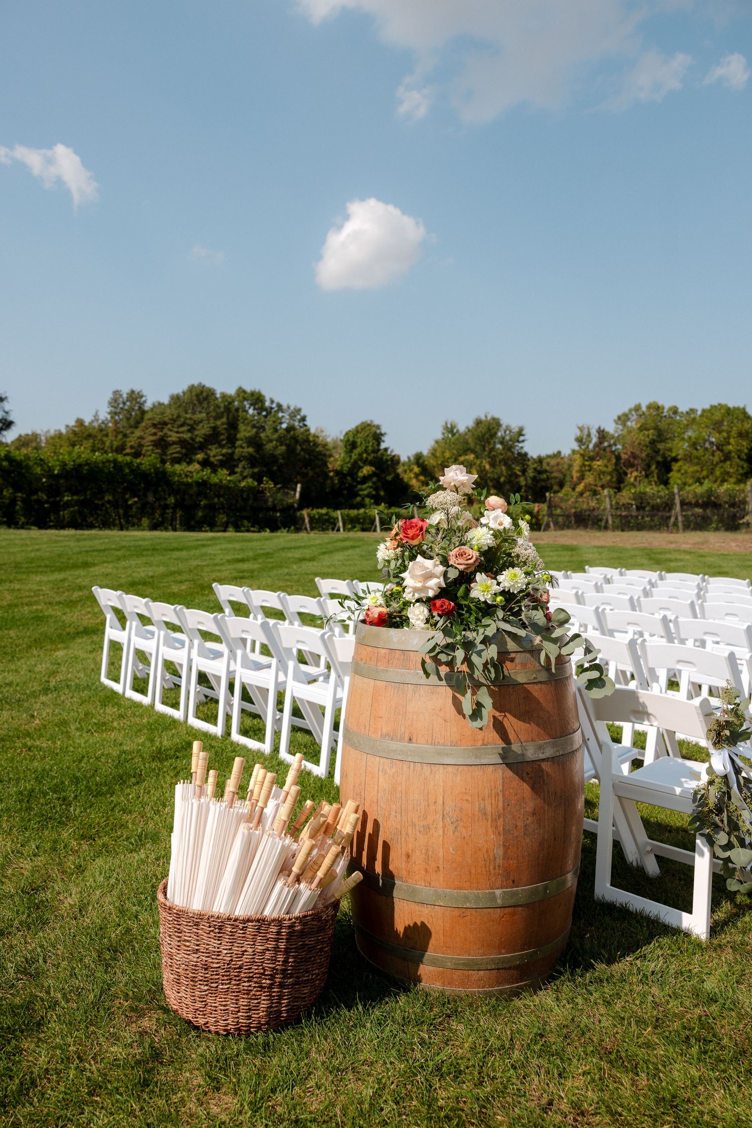 Outdoor wedding ceremony setup with white chairs, wooden barrel floral display, and basket of umbrellas on lush green lawn.