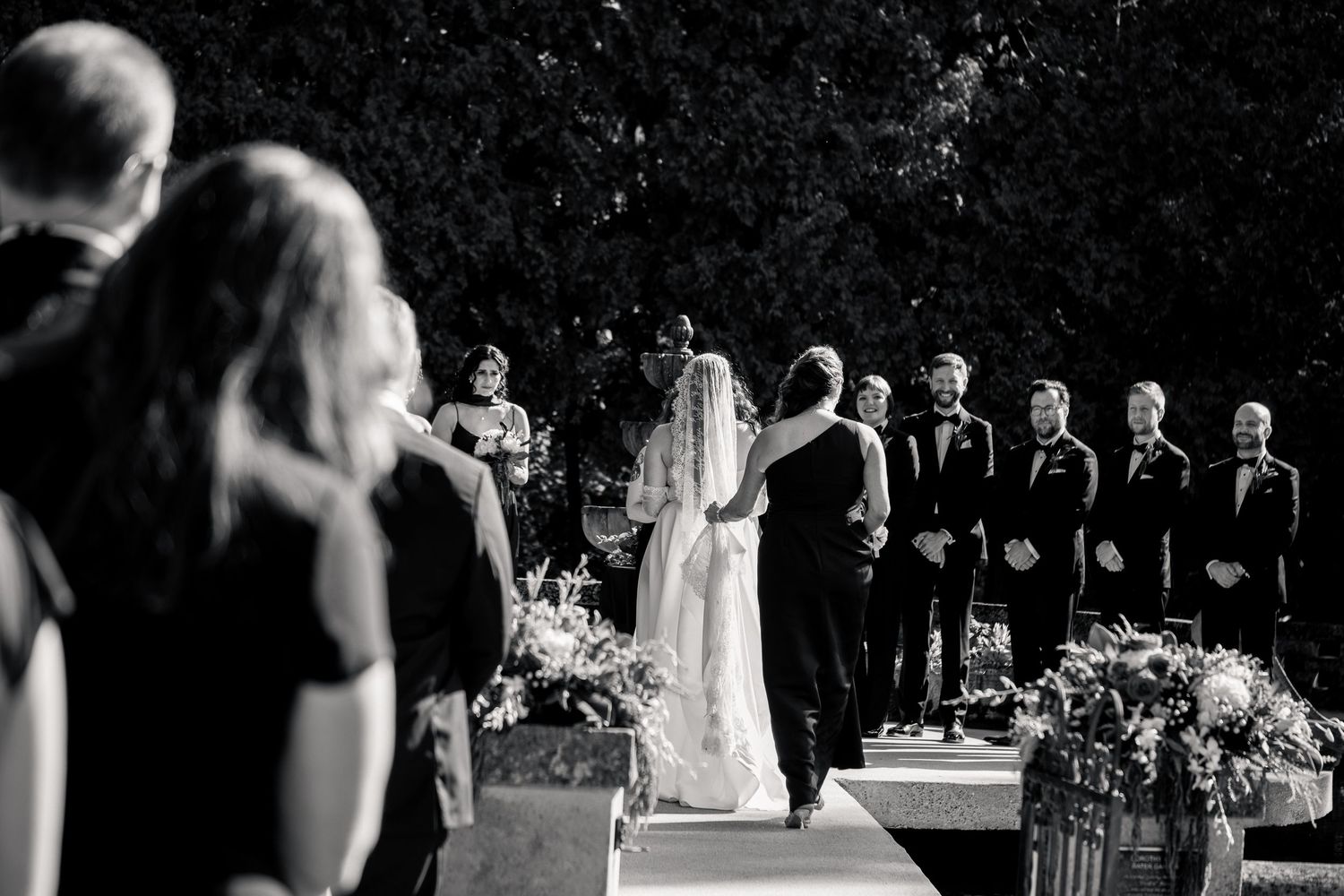 Black and white outdoor wedding ceremony with bride and groom at altar surrounded by wedding party and floral arrangements.