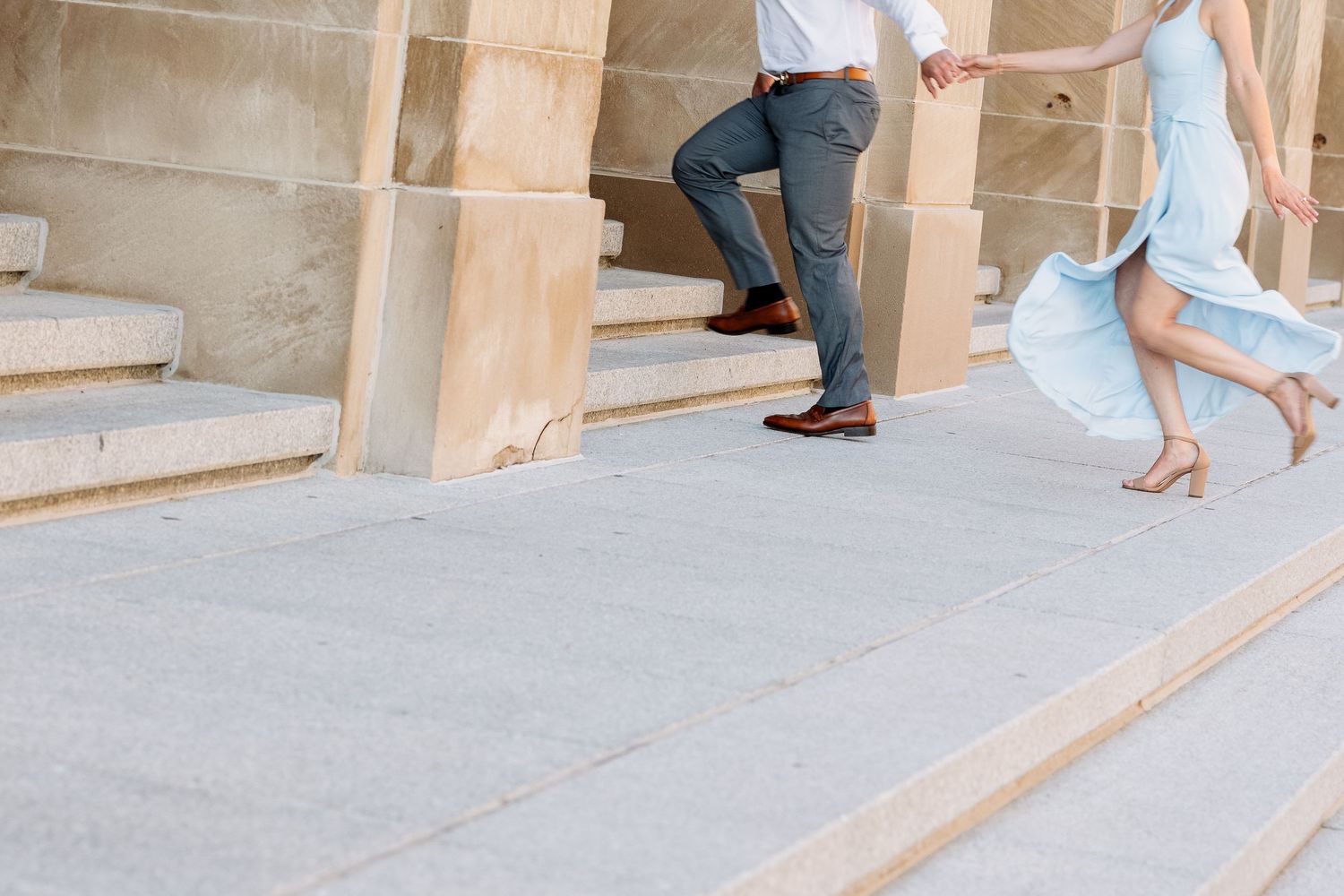 Couple walking up stone steps, man in blue pants and woman in flowing light blue dress, elegant outdoor setting.