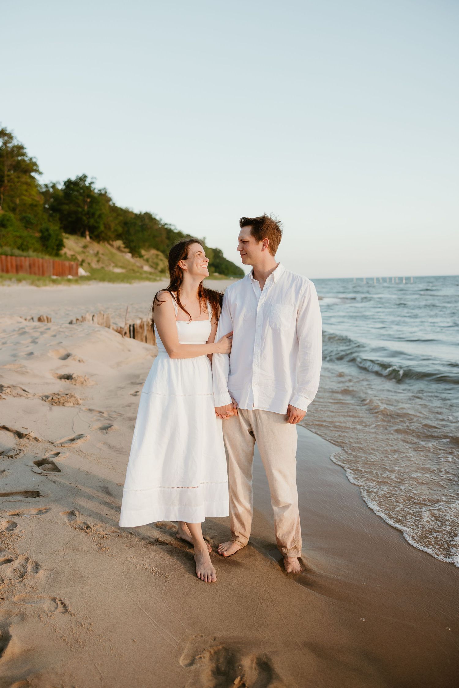 A couple walks barefoot on a sandy beach at sunset, wearing white outfits, smiling at each other near the ocean.