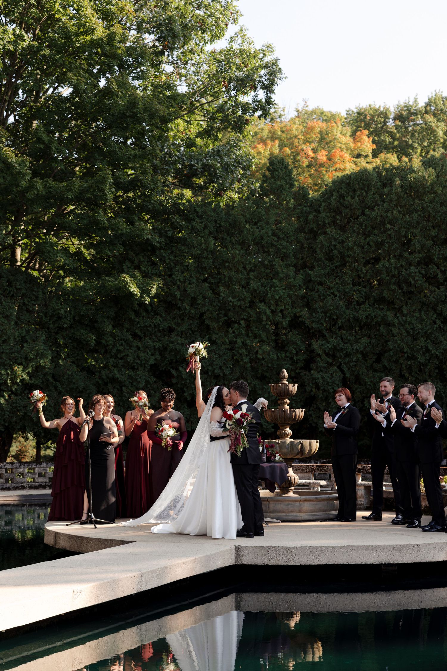 Bride and groom share first kiss on outdoor wedding ceremony stage surrounded by bridesmaids in burgundy and groomsmen.