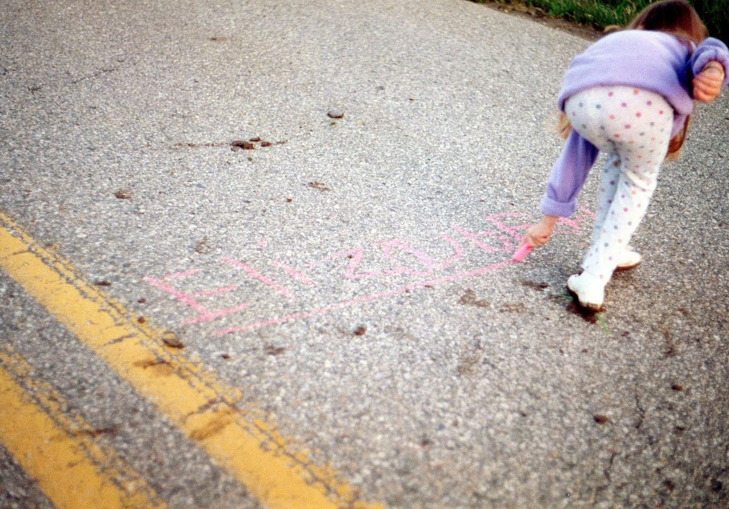 Young child drawing pink chalk art on a road near a yellow line outdoors.