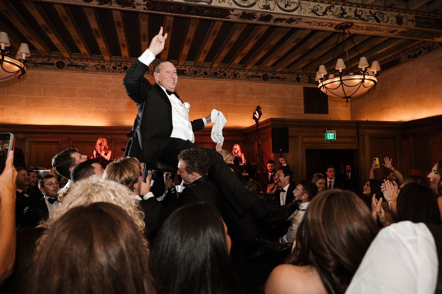 Man in tuxedo raised on shoulders by crowd waving napkin at wedding celebration.