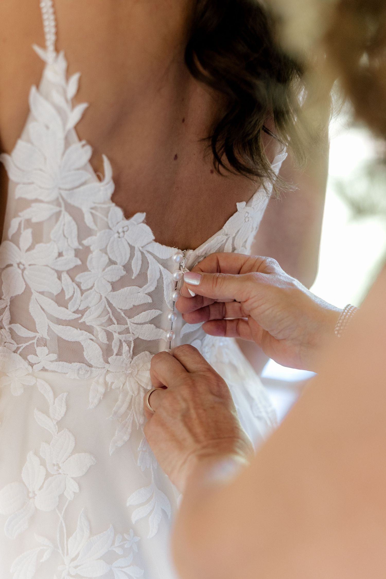 Close-up of hands fastening buttons on a floral lace wedding dress during bridal preparations.
