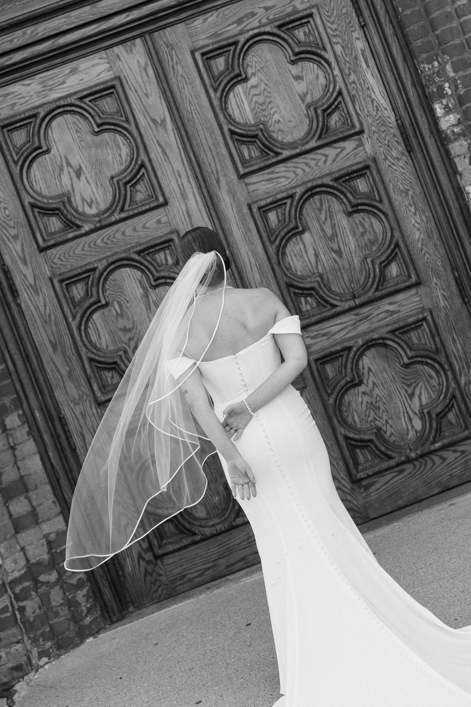 Black and white photo of a bride in a fitted off-shoulder gown and veil standing before ornate wooden church doors.