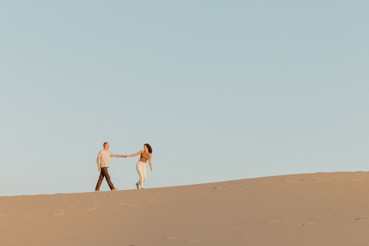 Couple holding hands walking on sand dunes under clear blue sky.