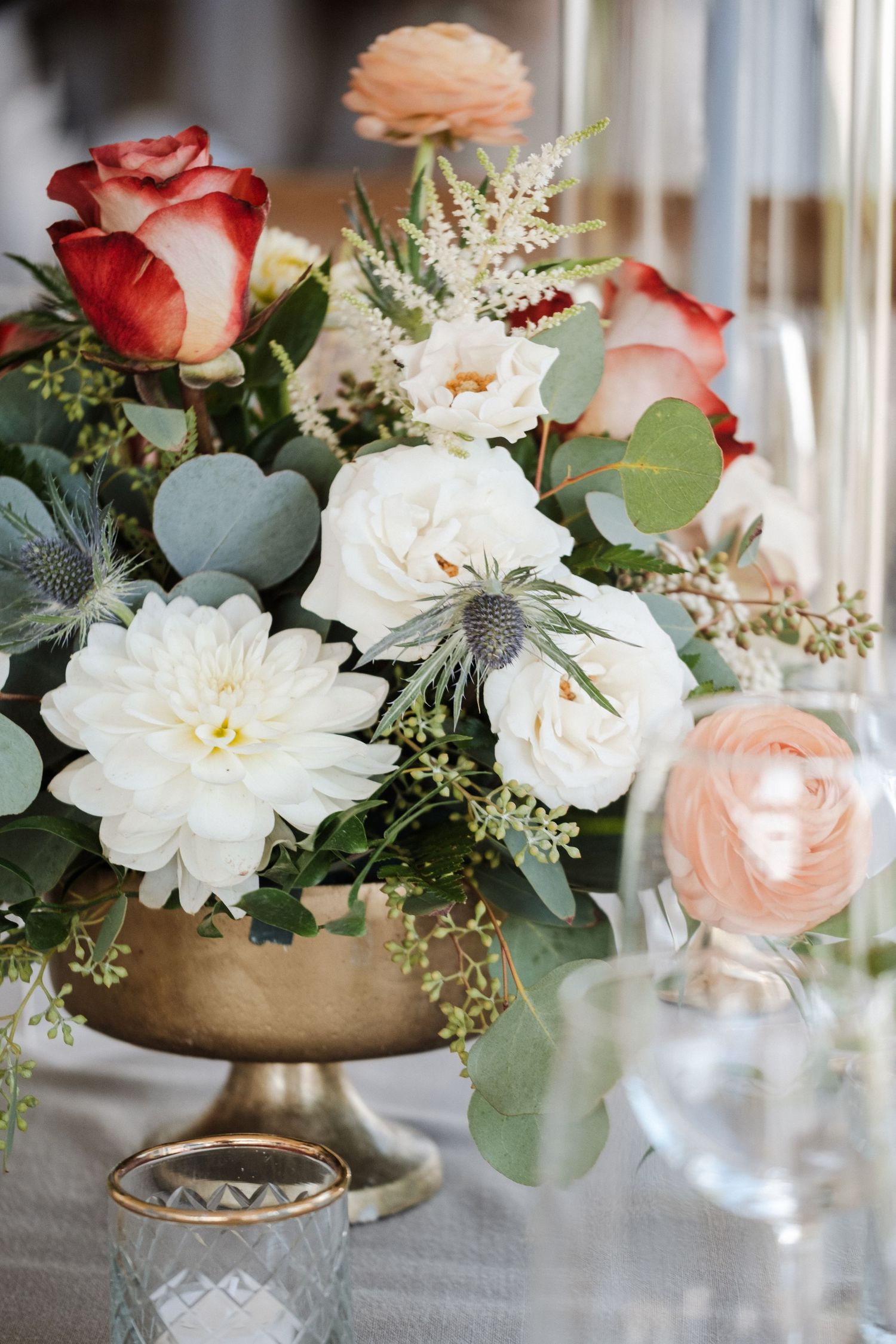 Elegant wedding centerpiece with white dahlias, red roses, peach ranunculus, and eucalyptus in gold vase.