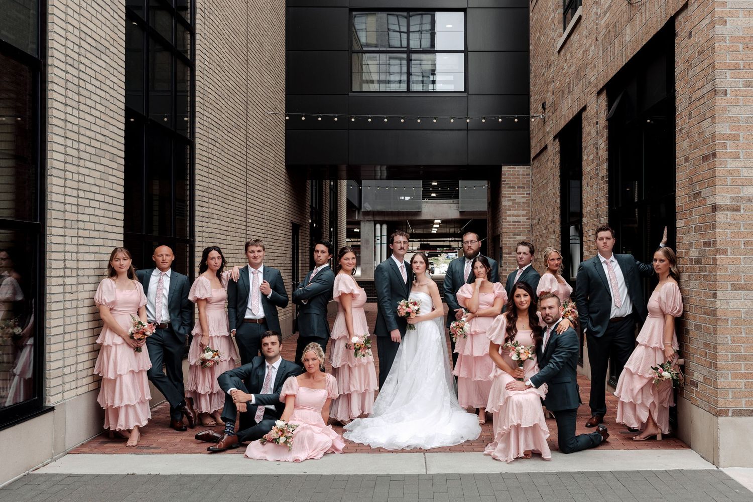 Large wedding party poses outside brick building, bridesmaids in pink tiered dresses with groomsmen in dark suits.