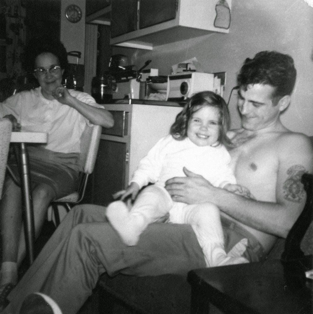 Vintage black and white family photo of a man holding a young girl while a woman sits nearby in a kitchen.