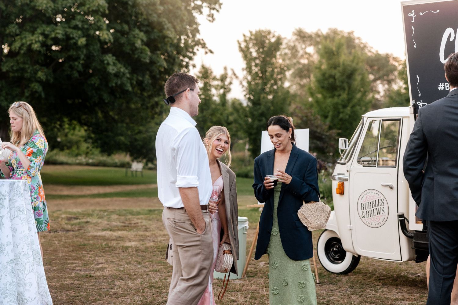 Wedding guests mingling outdoors at golden hour near vintage white food truck at reception.