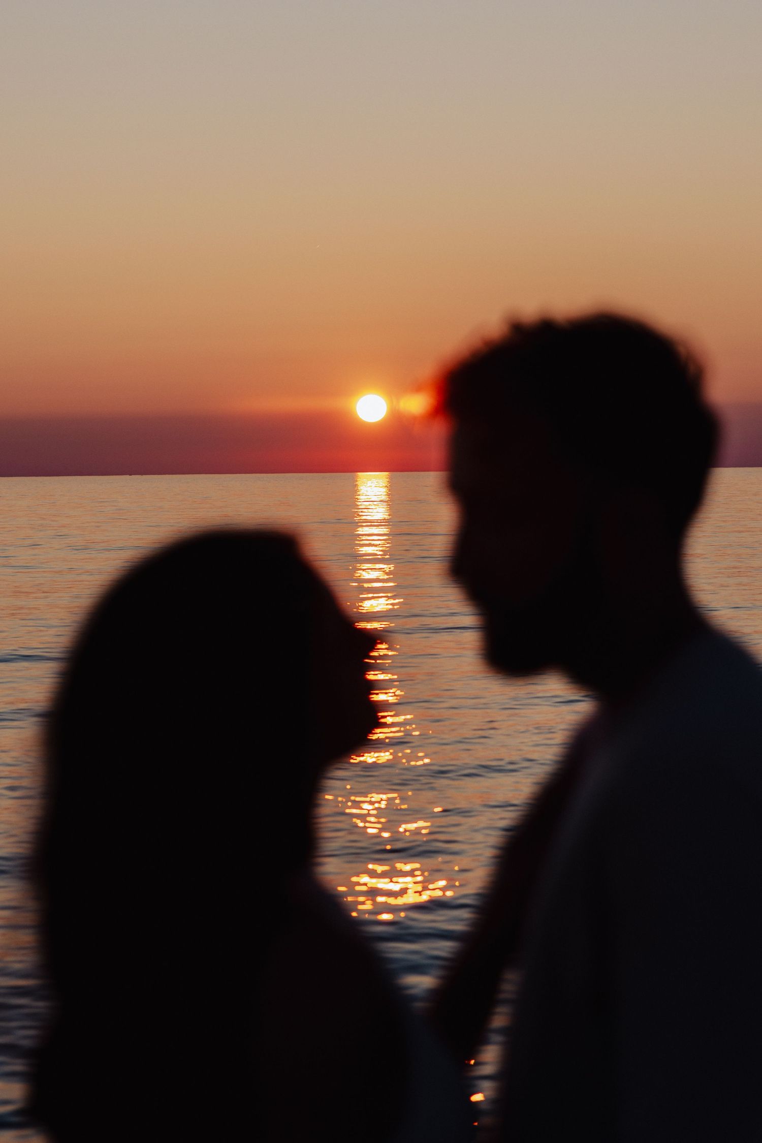 Romantic silhouette of a couple facing each other against a vibrant orange sunset over calm ocean waters.