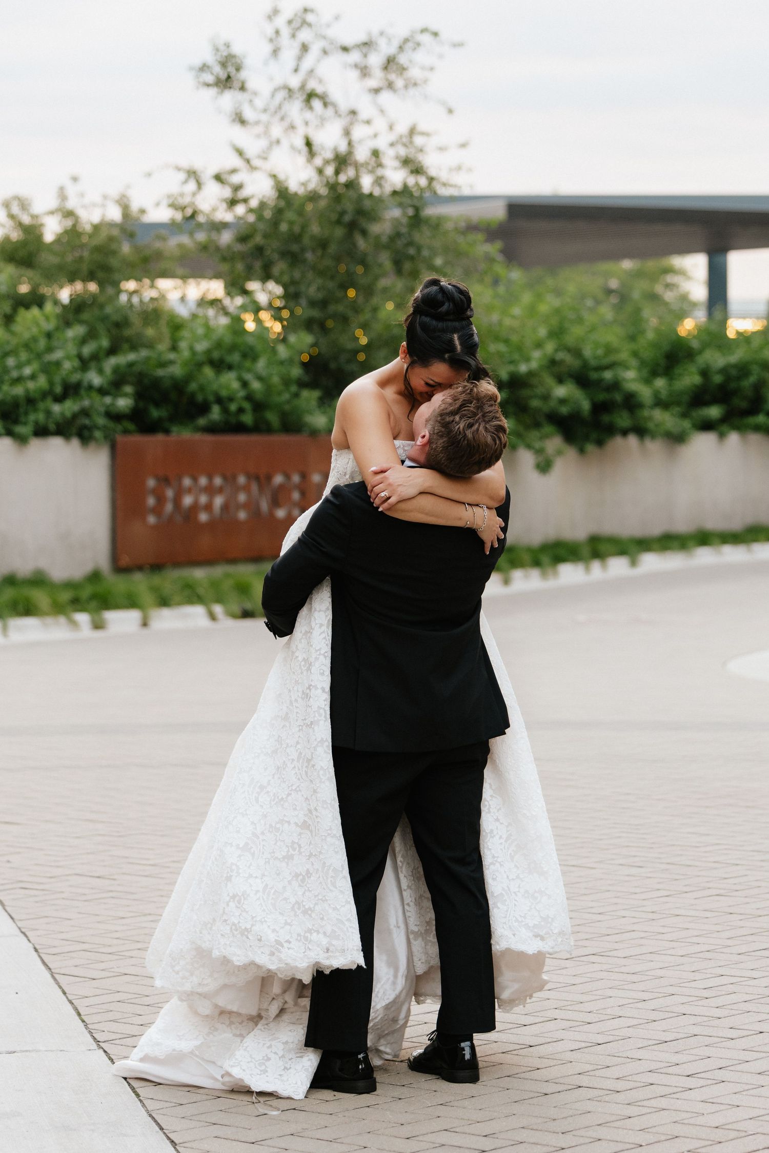 Groom lifts bride in white lace wedding gown outdoors, sharing a romantic embrace on their wedding day.
