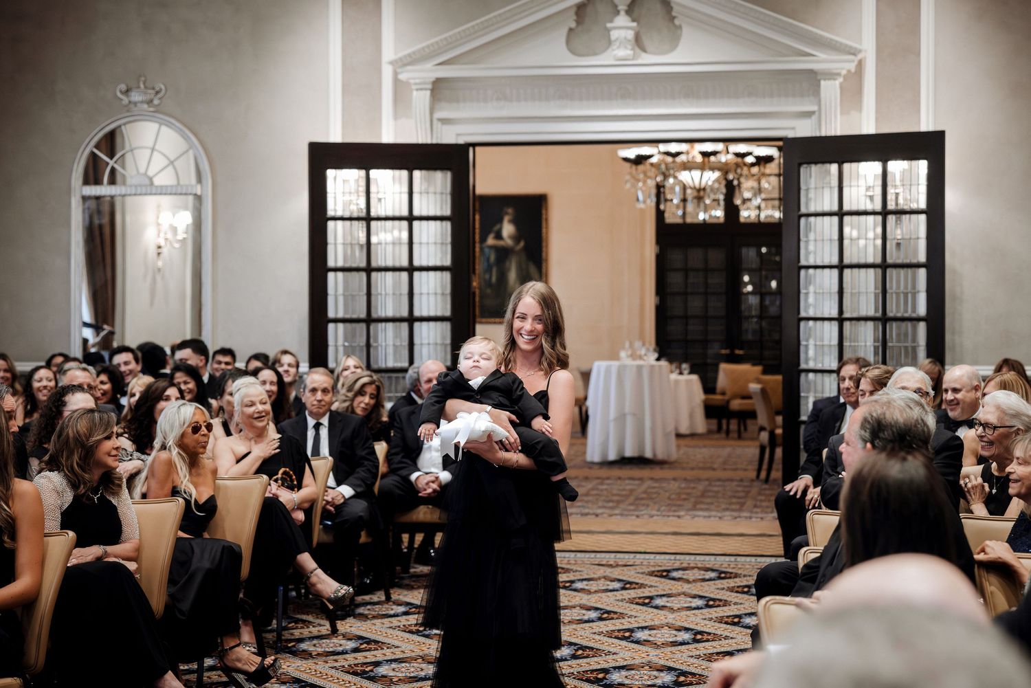 Woman carries a baby down the wedding ceremony aisle in a grand ballroom filled with seated guests in black attire.