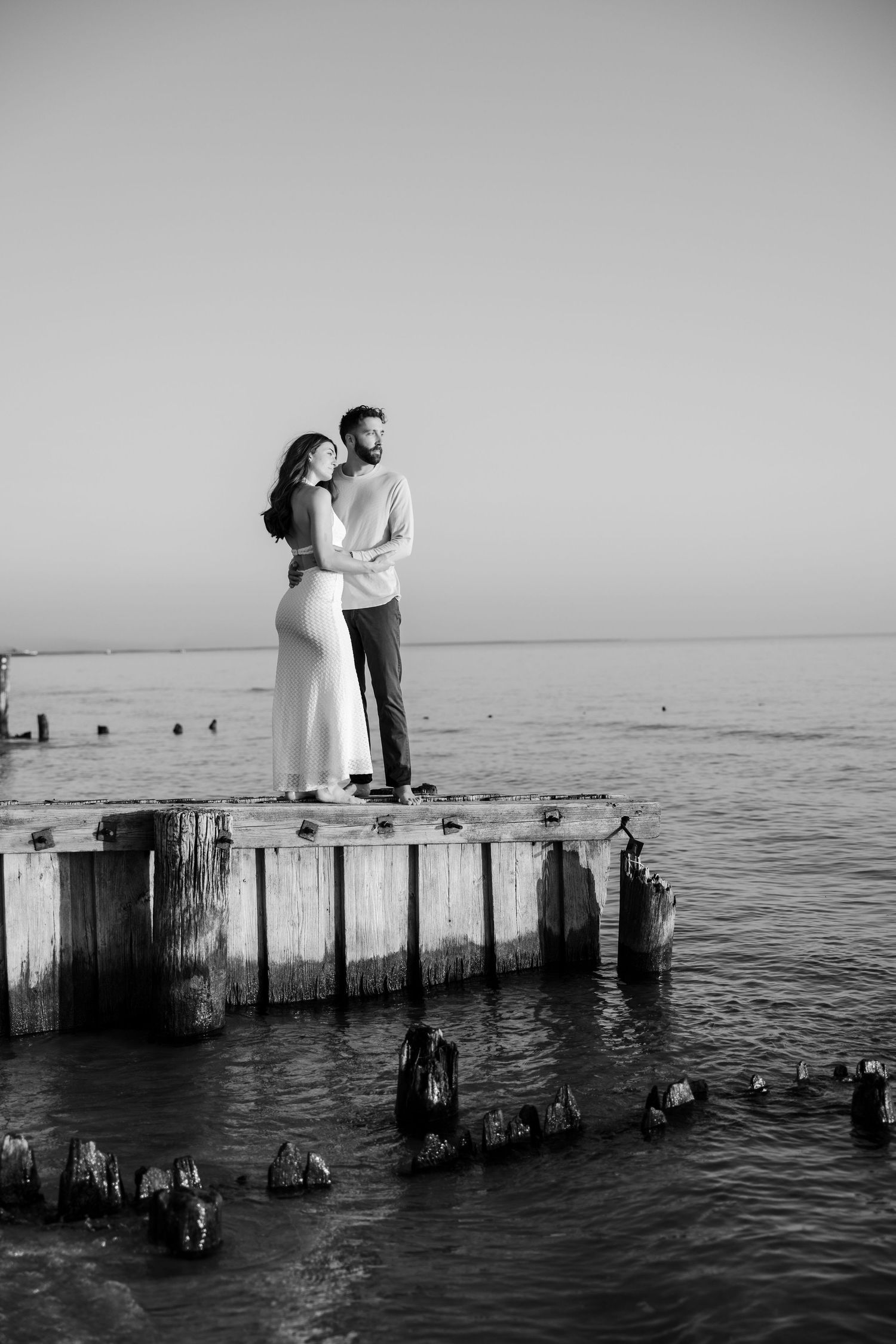 Black and white photo of a couple embracing on a weathered pier over calm ocean waters at sunset.