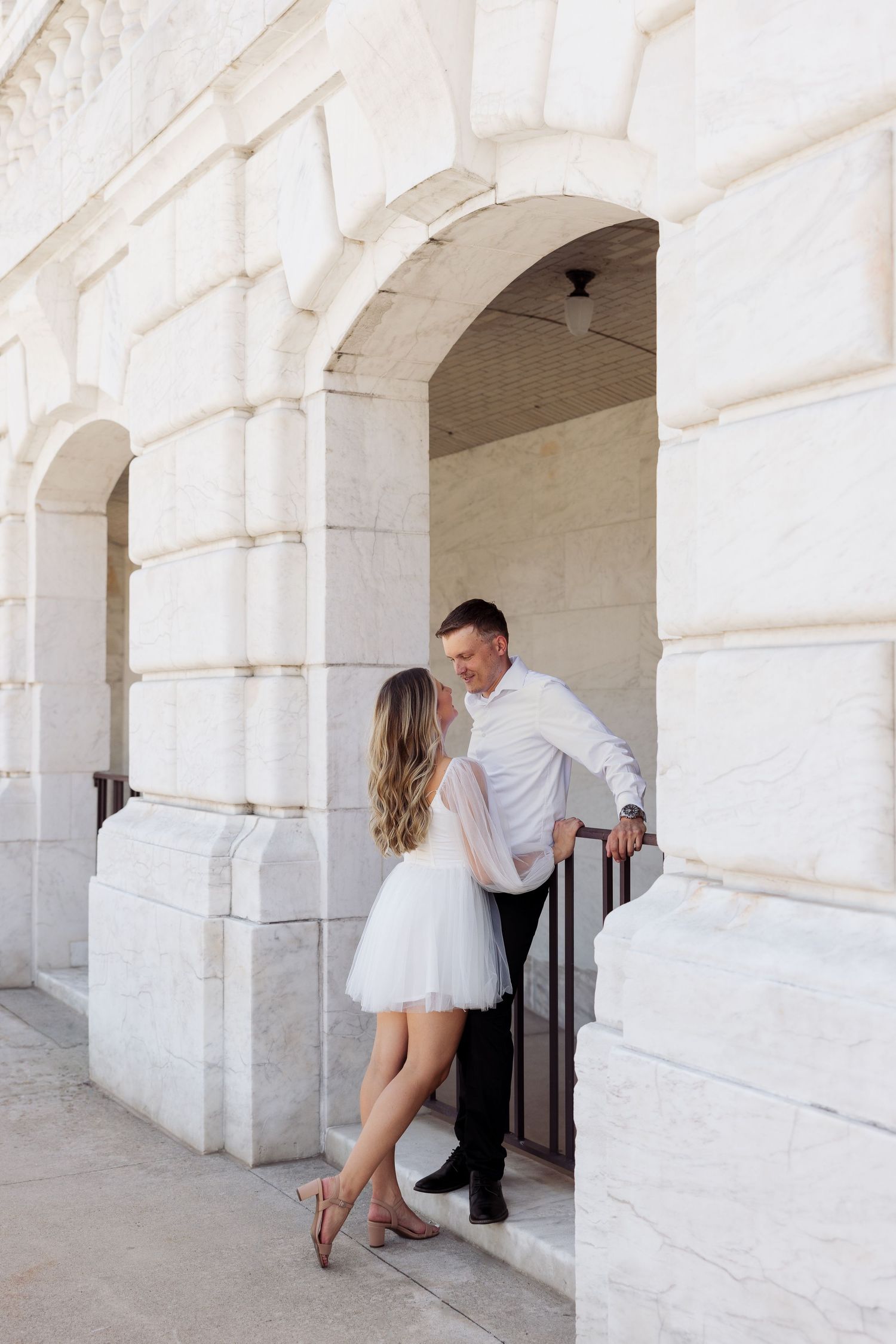 Couple sharing an intimate moment under a white stone archway of a classic architectural building.