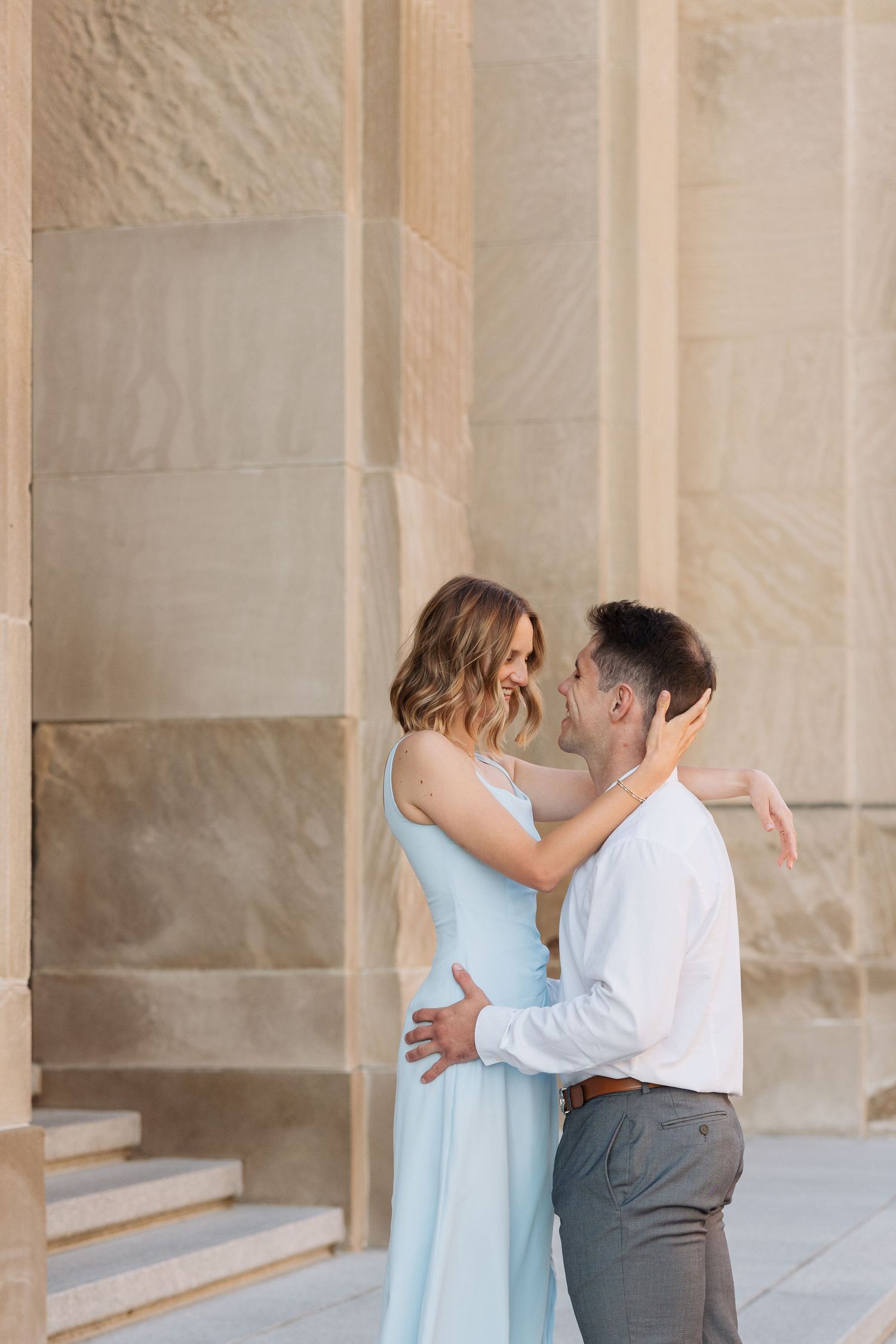 Romantic couple hugging between beige stone pillars, woman in pale blue dress touching mans face.