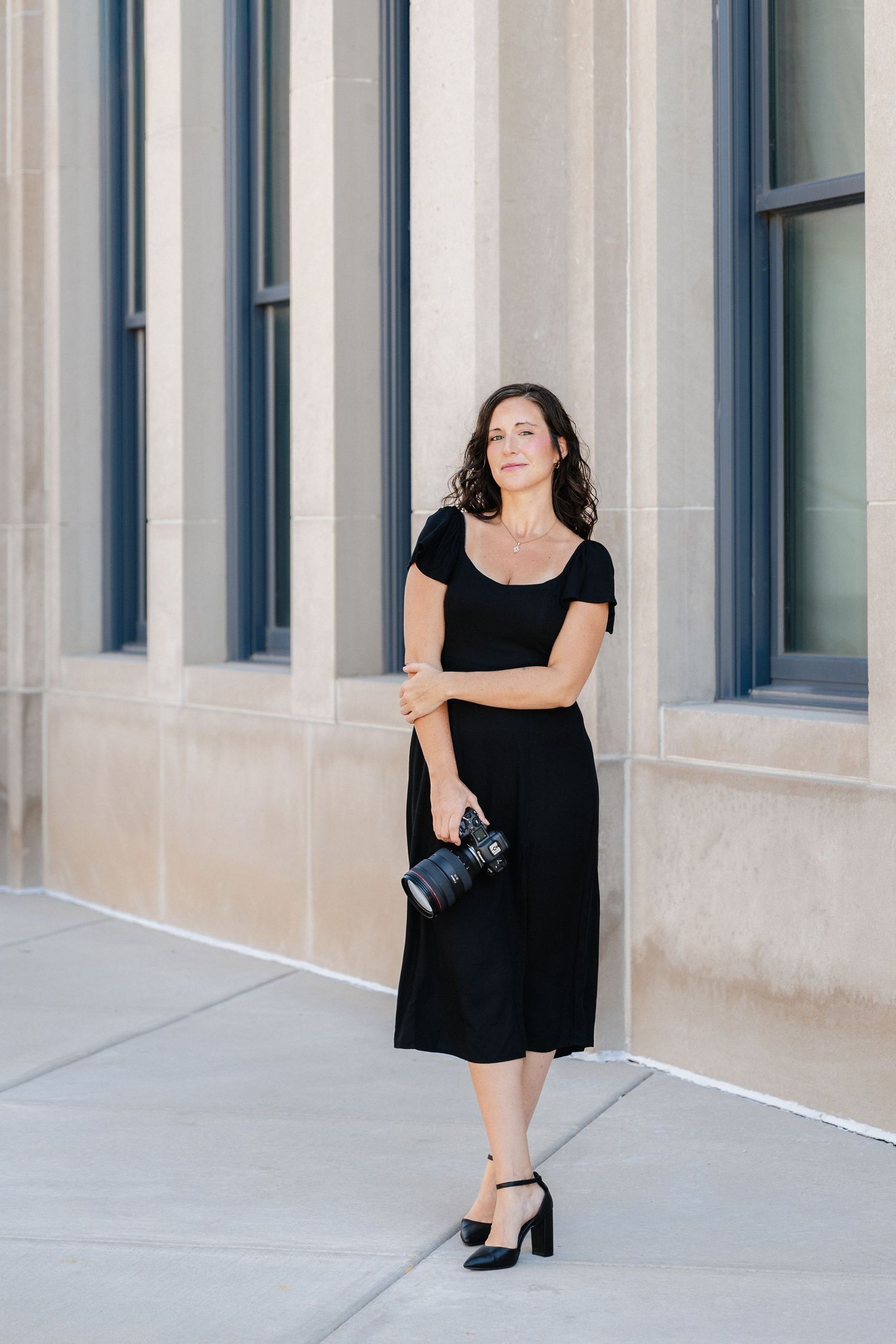 Woman in black dress holding camera stands confidently outside stone building.