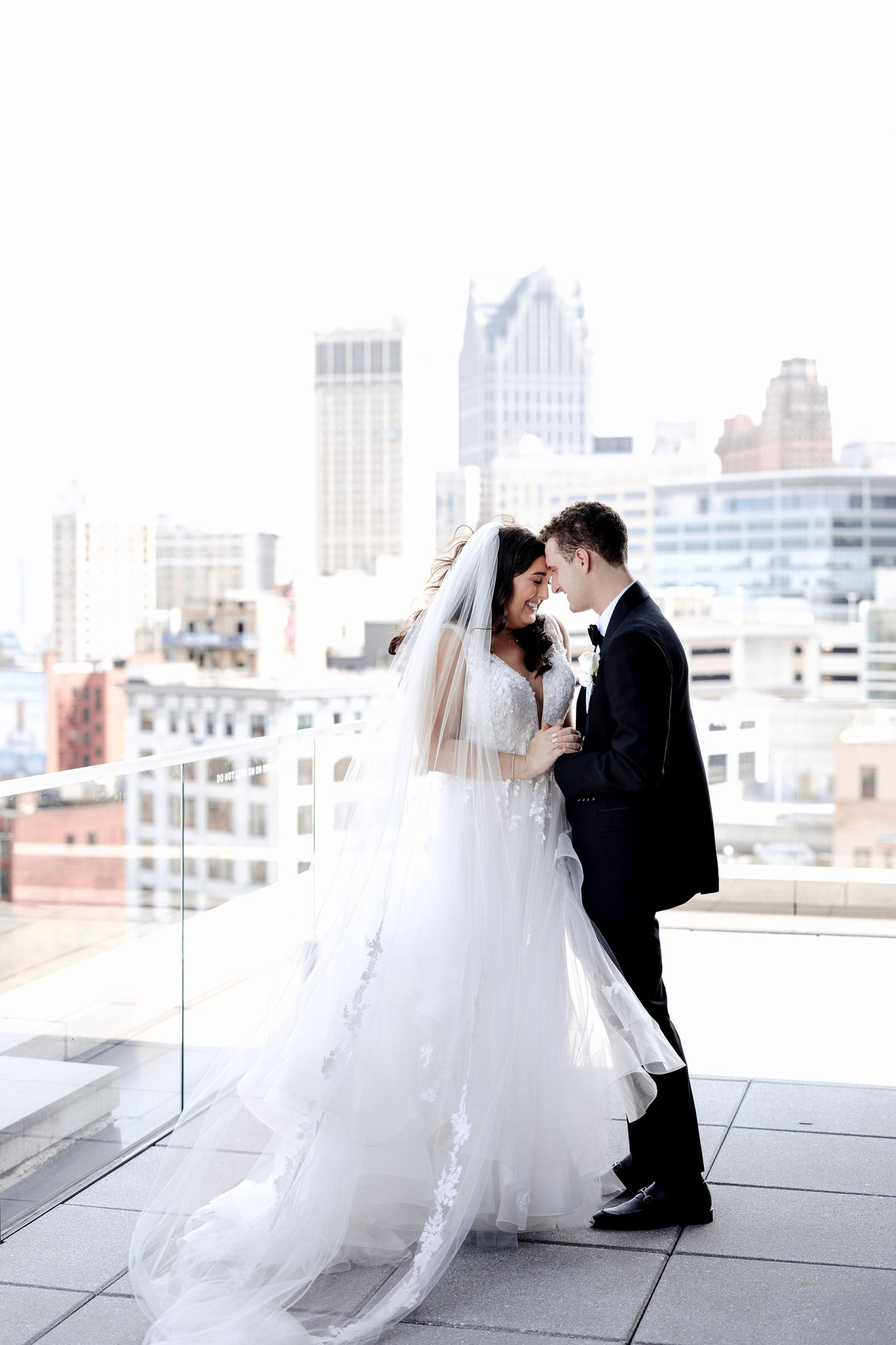 Bride and groom nearly kiss on a sunlit rooftop, her flowing white tulle gown and veil billowing in the breeze.