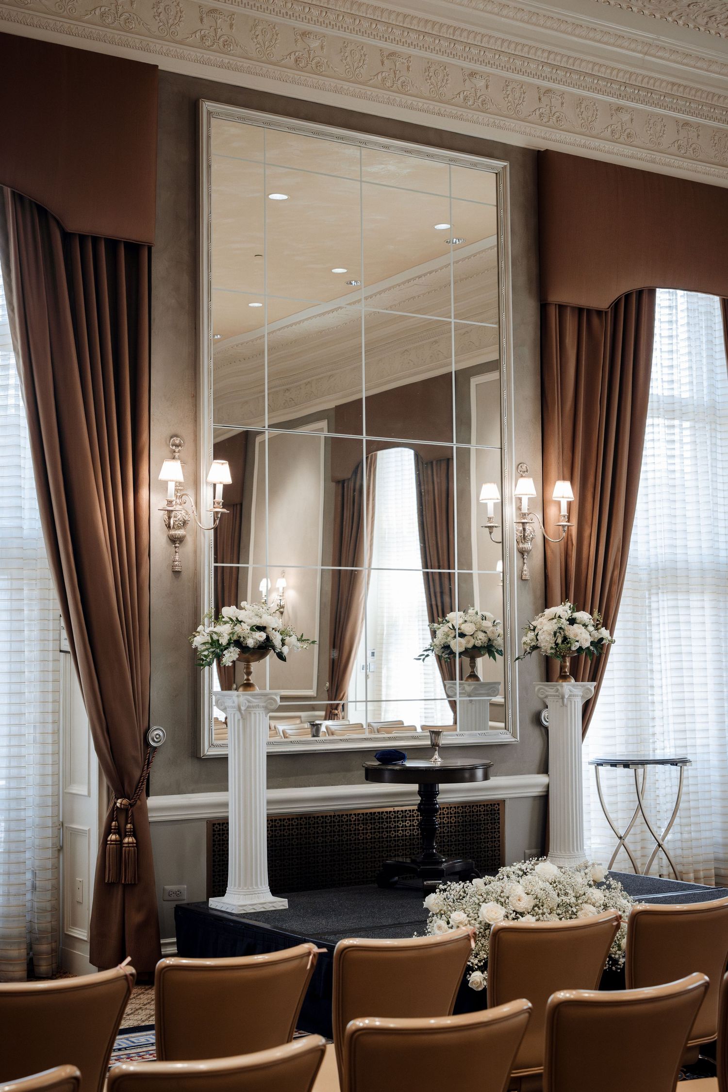 Elegant wedding ceremony altar with large ornate mirror, white flower arrangements on white pedestals, and tan chairs.