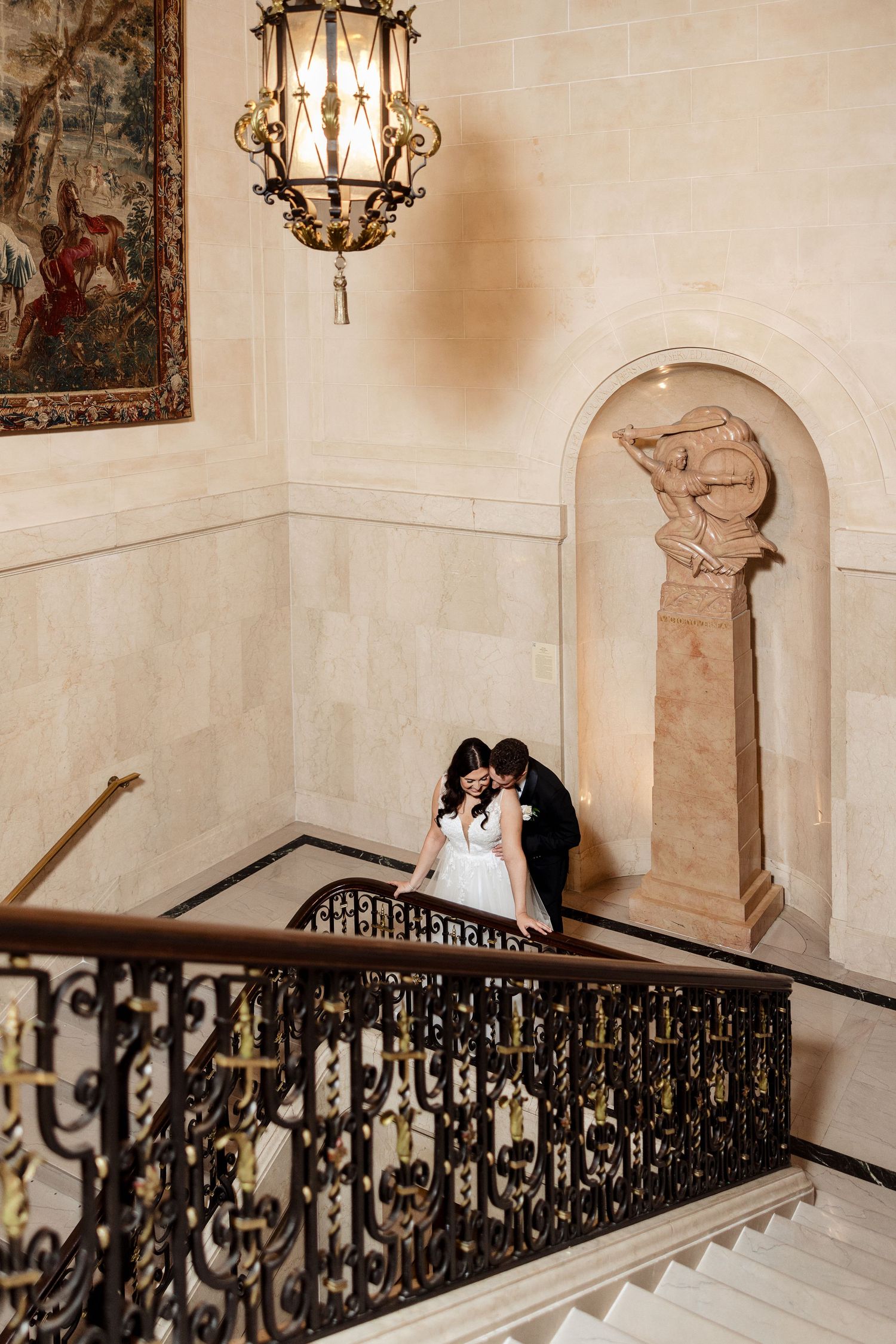 Bride and groom kissing on an elegant staircase with wrought iron railings and a chandelier above.