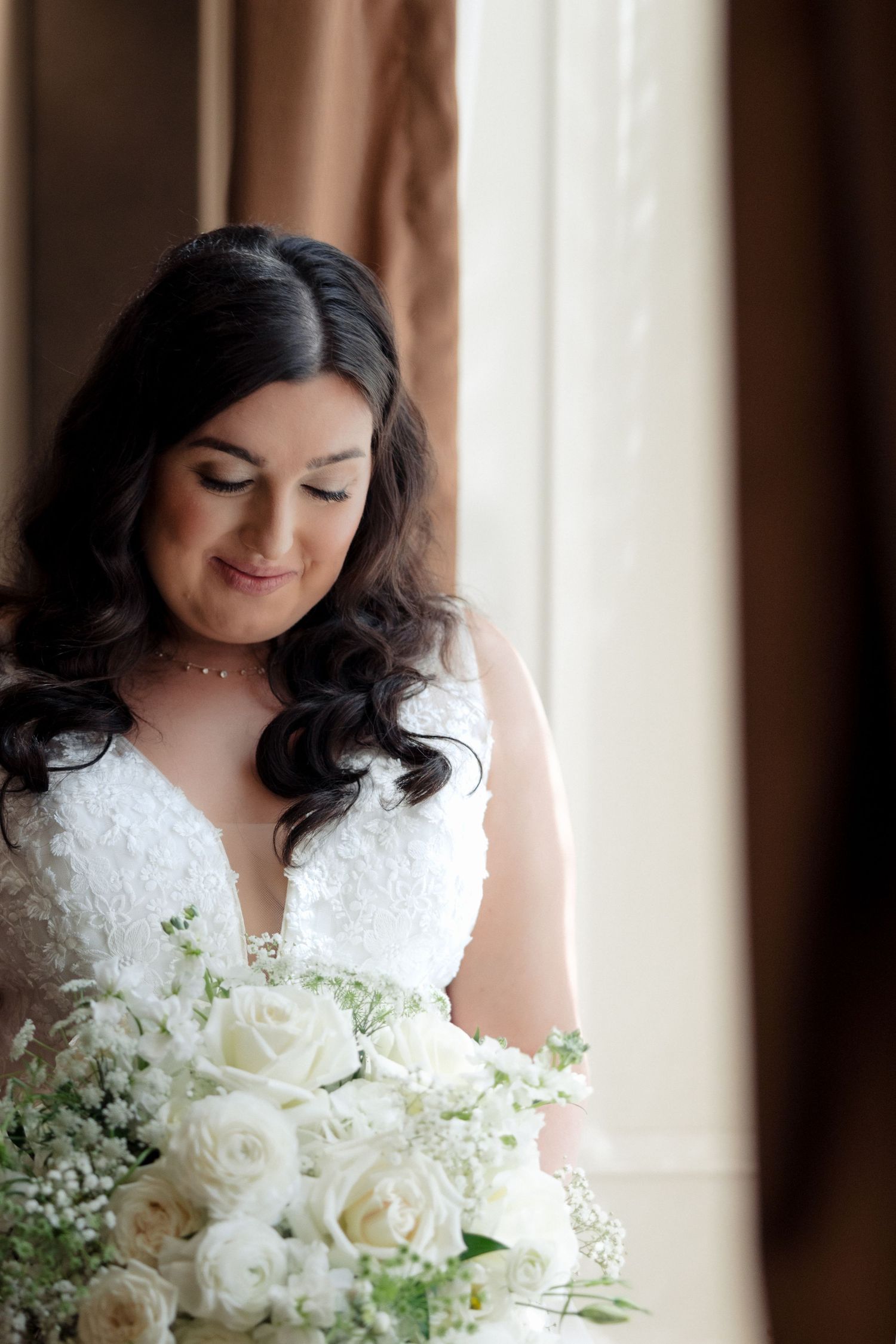 Bride in white lace dress looks down at white rose bouquet by window.