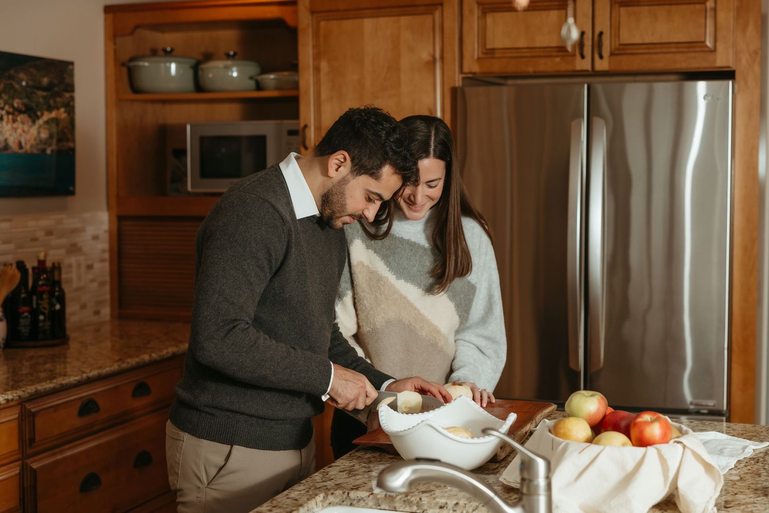 A couple smiles together while cooking in a warm wooden kitchen with fresh apples on the counter.