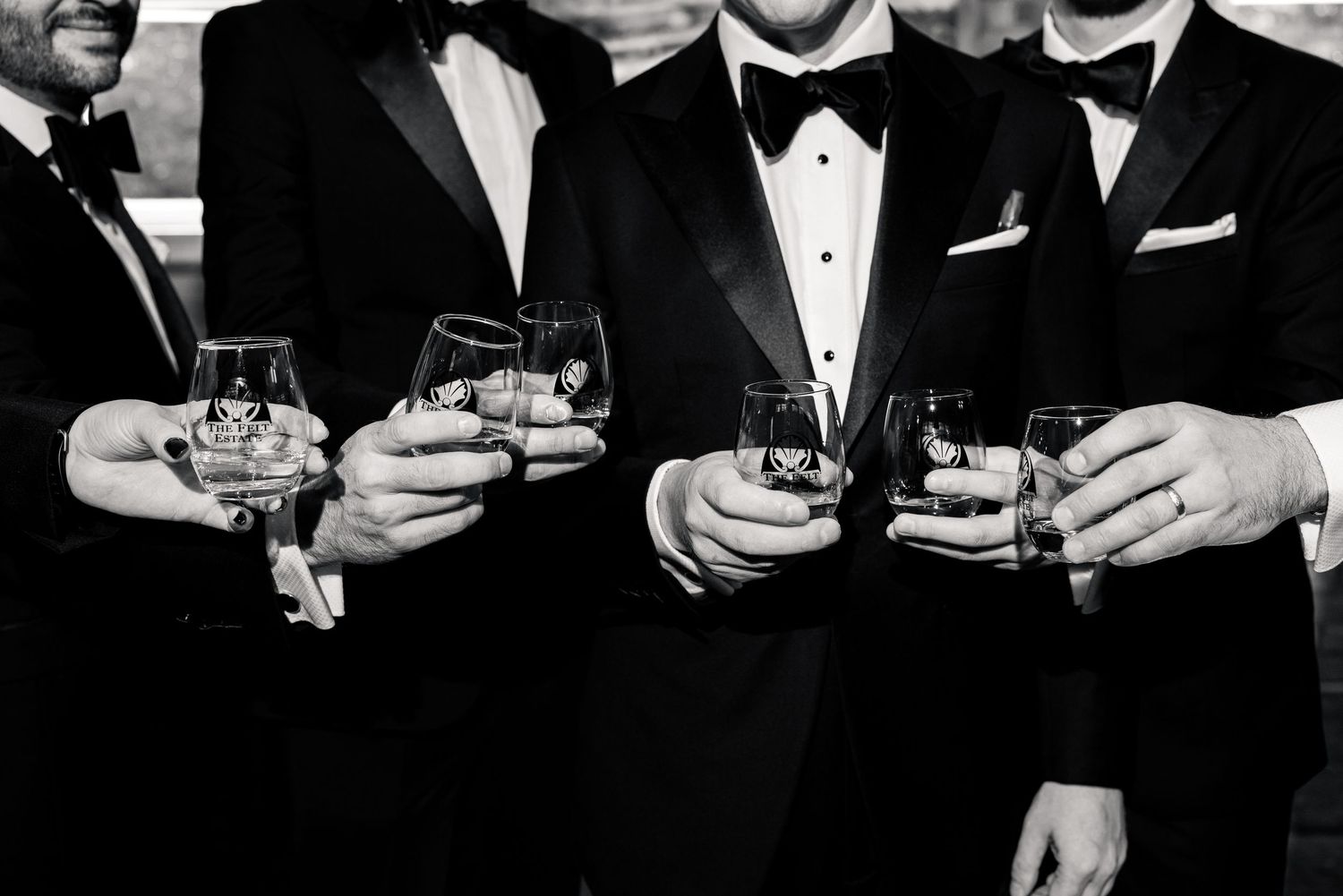 Black and white close-up of groomsmen in tuxedos holding engraved whiskey glasses at a wedding.