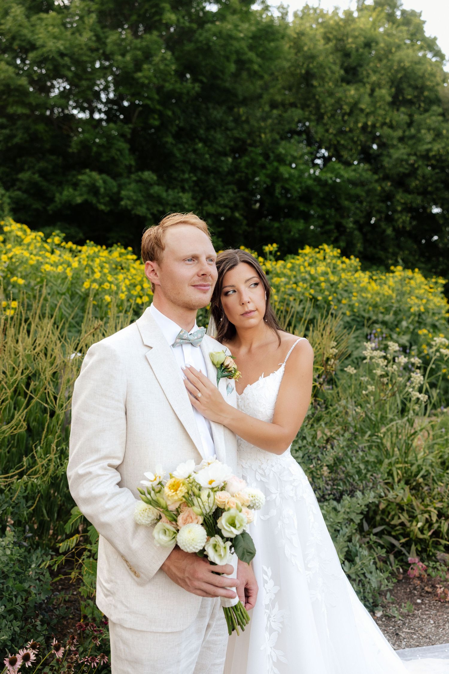 Bride and groom pose in garden with yellow flowers, groom in cream suit holding bouquet, bride in white lace gown.